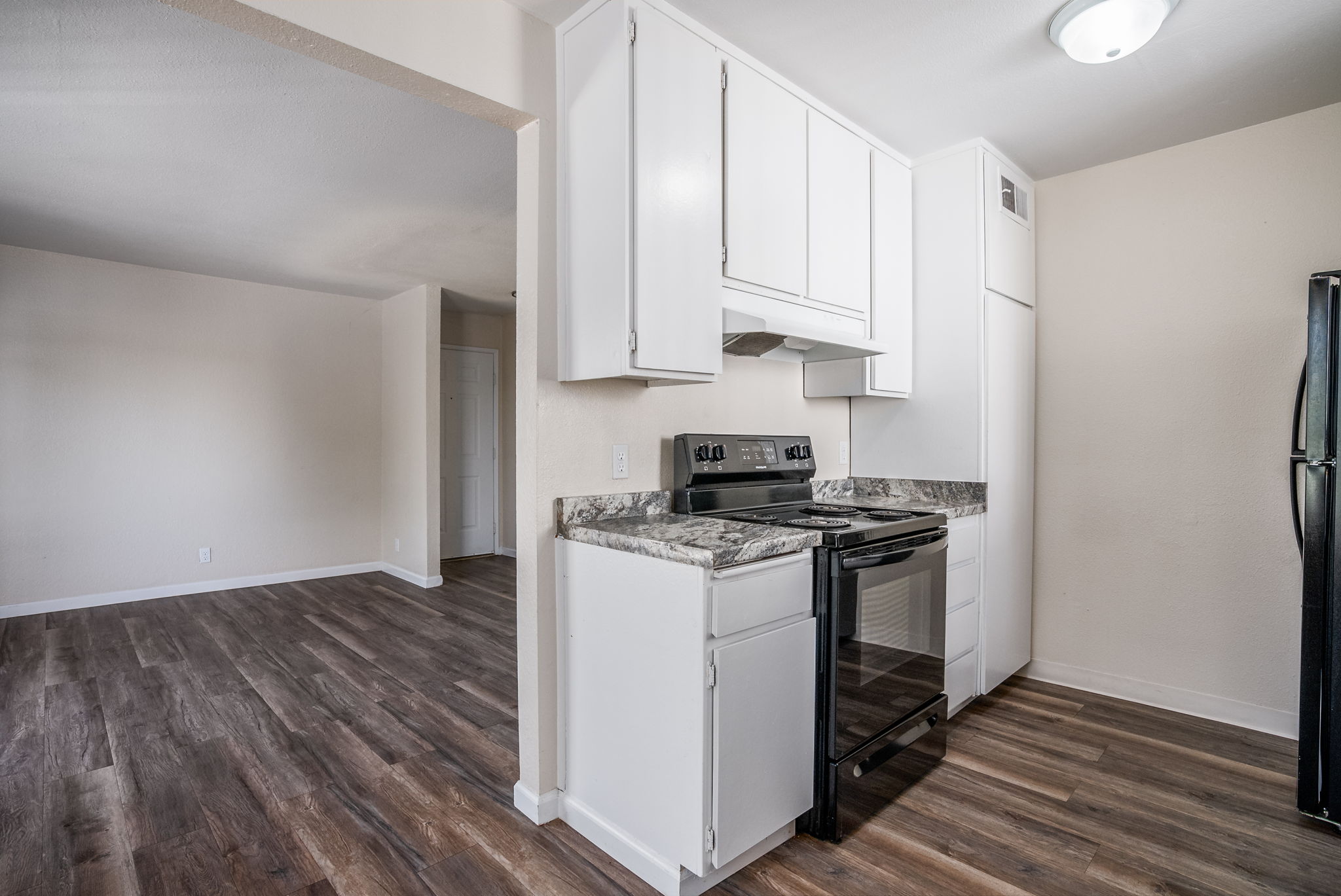A clean, modern kitchen with white cabinetry, a black stove, and granite countertops. The kitchen opens to a spacious living area with light-colored walls and hardwood flooring. Bright lighting fixtures enhance the inviting atmosphere.