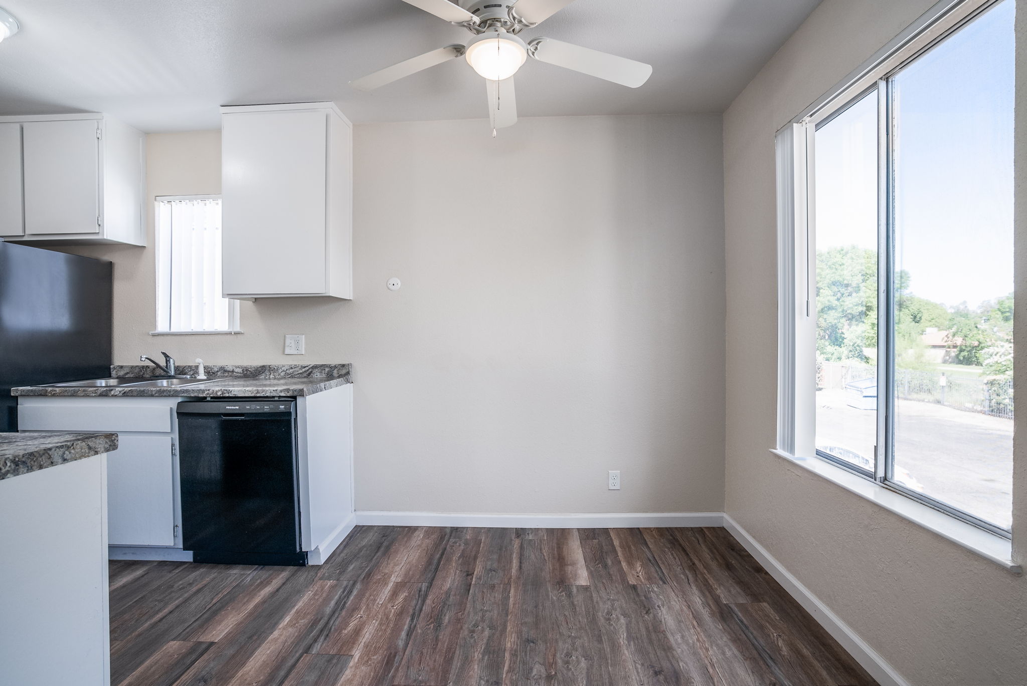 A bright kitchen area featuring white cabinets, a black refrigerator, and a window that lets in natural light. The floor is made of wood-like laminate. A ceiling fan is installed above, adding to the open and airy feel of the space. The walls are painted in a light color, enhancing the room's brightness.