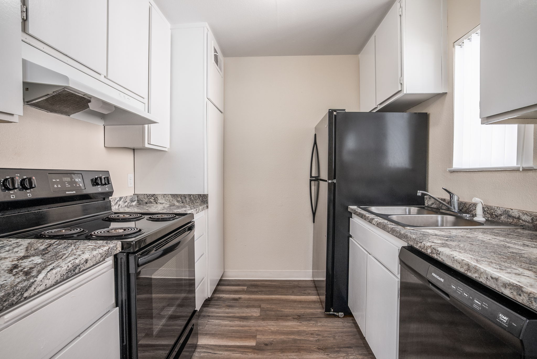 A modern kitchen featuring white cabinets, granite countertops, and stainless steel appliances. The layout includes a black refrigerator, an electric stove with an overhead hood, a double sink, and a dishwasher. The flooring is dark wood, and natural light enters through a window.