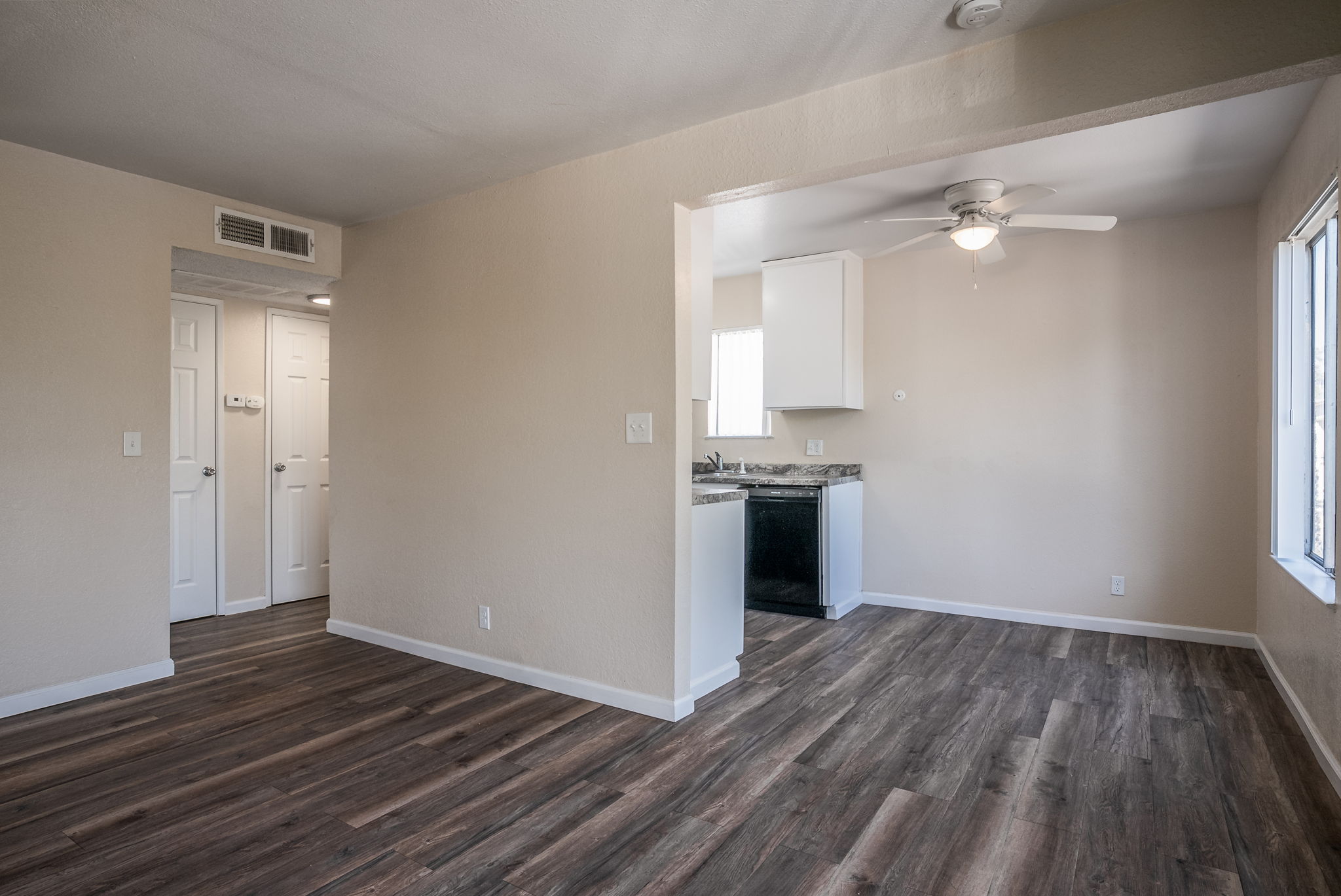 A bright, unfurnished living space with light-colored walls and wood-style flooring. There is a ceiling fan, a window providing natural light, and an open view into a small kitchen area with a countertop, cabinets, and appliances. A doorway leads to another room, indicating a cozy apartment layout.