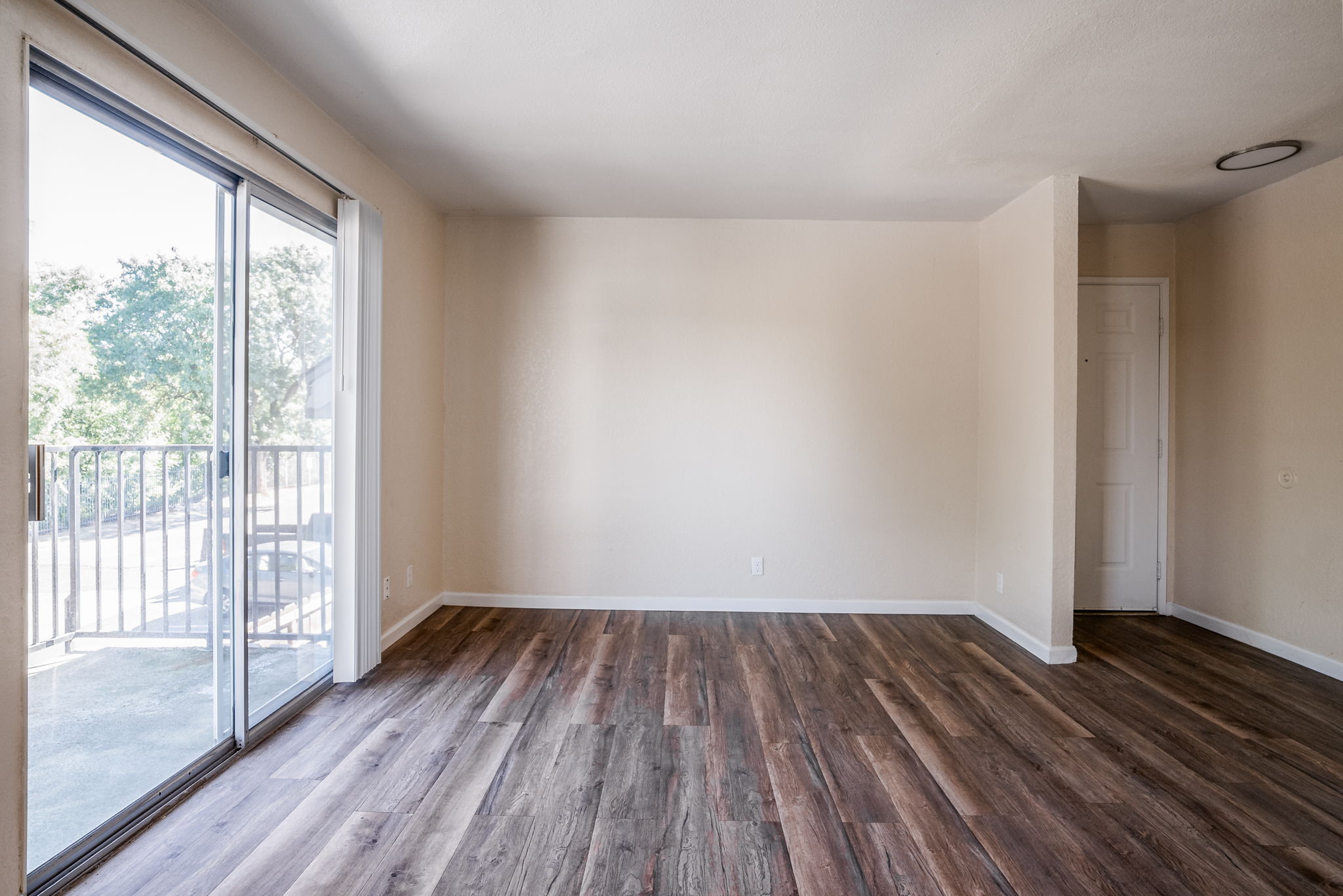 Empty room featuring light-colored walls, large sliding glass door with a view of greenery outside, and wooden laminate flooring. The space is well-lit and has a minimalistic appearance, with a corner door visible in the background.