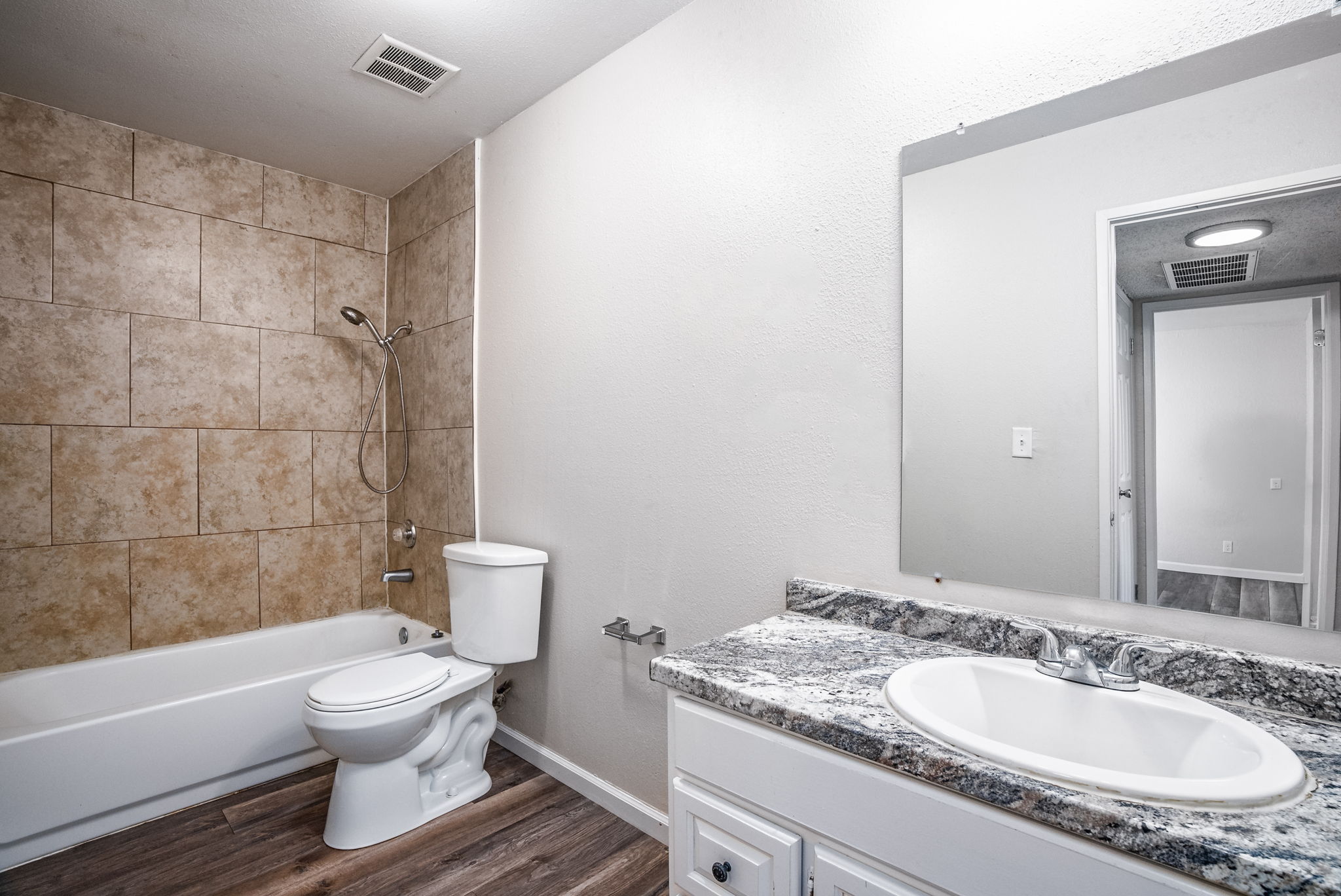 A clean and modern bathroom featuring a white bathtub with a showerhead, a toilet, and a vanity with a granite countertop and sink. The walls are tiled with light-colored ceramic tiles, and the flooring is a wood-like laminate. A mirror above the sink reflects the bright, spacious layout.