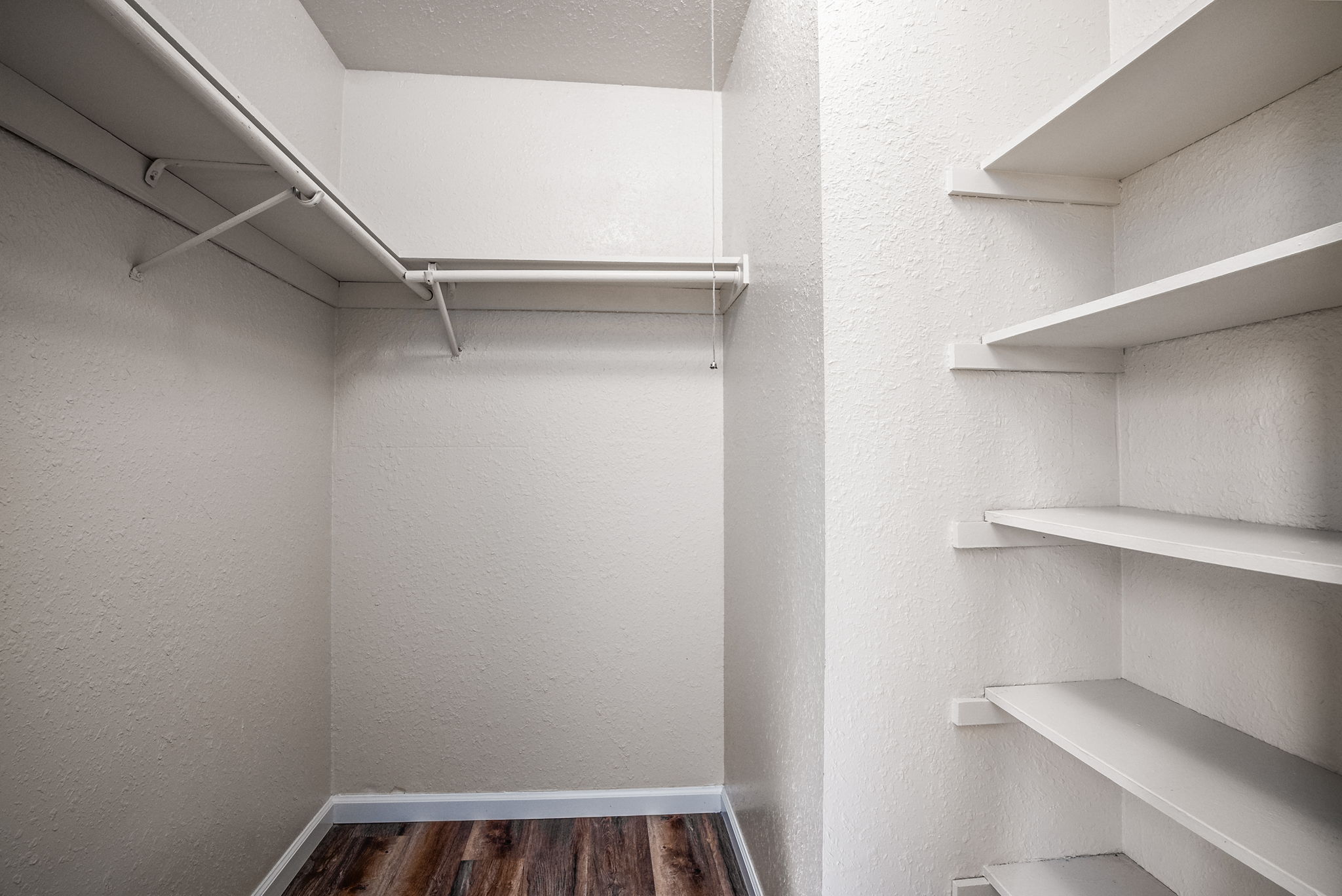 Interior of a small, empty closet featuring white walls, light wooden flooring, a hanging rod on the left, and open shelves on the right. The space appears clean and organized, with no items stored inside.