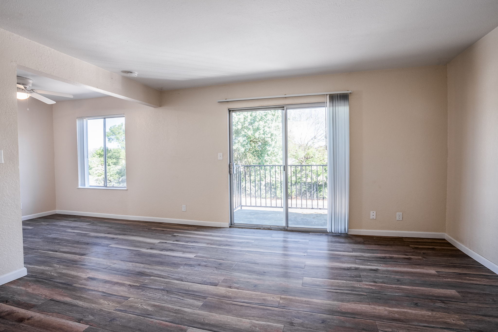 Spacious, empty living room with light-colored walls and brown wooden flooring. Features a large window letting in natural light, and a sliding glass door leading to a balcony with views of greenery. The room is well-lit, creating an inviting atmosphere.