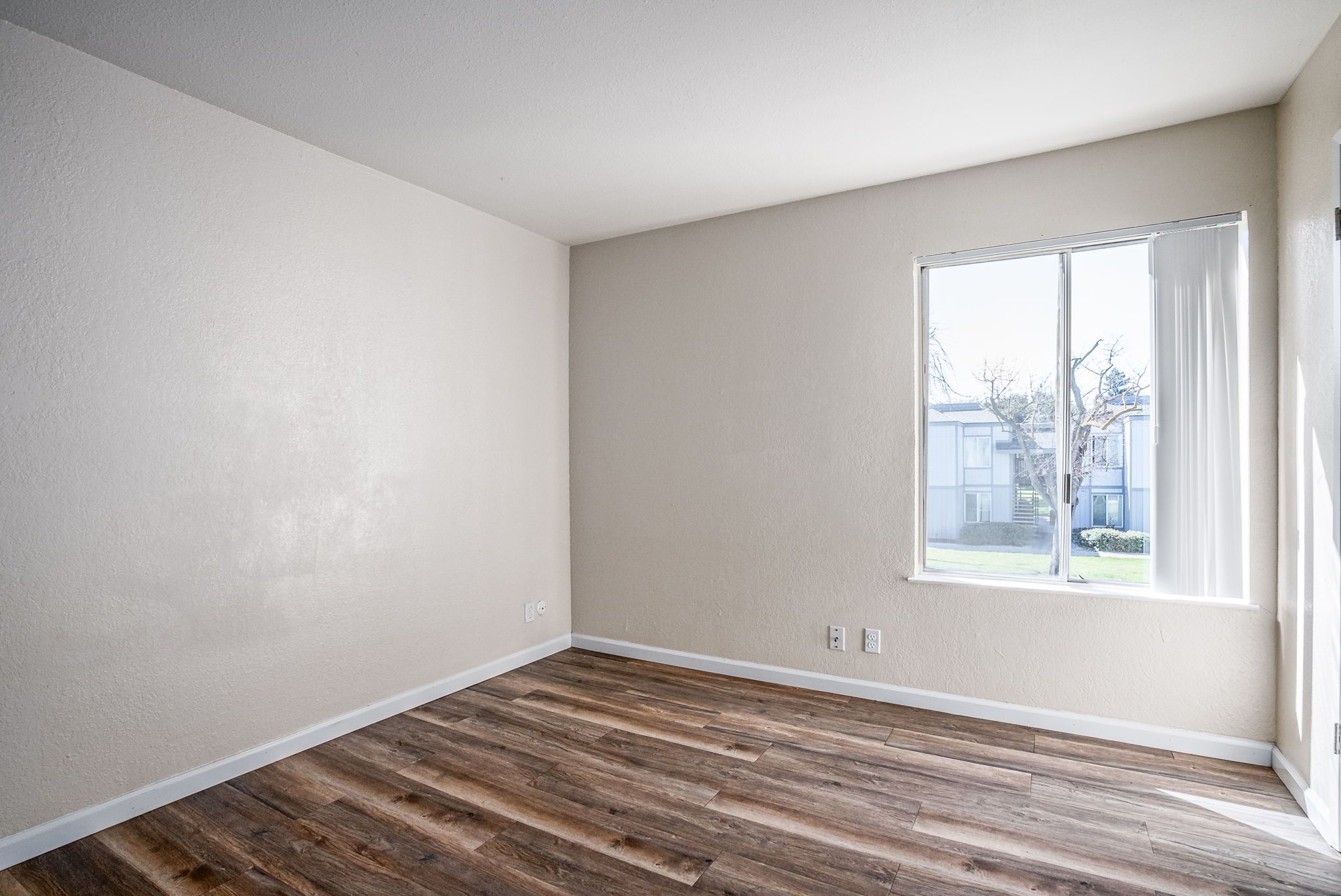A vacant room with light-colored walls and brown wooden flooring. A large window lets in natural light, showing a view of a green outdoor area. The space is minimalistic, with no furniture or decorations.