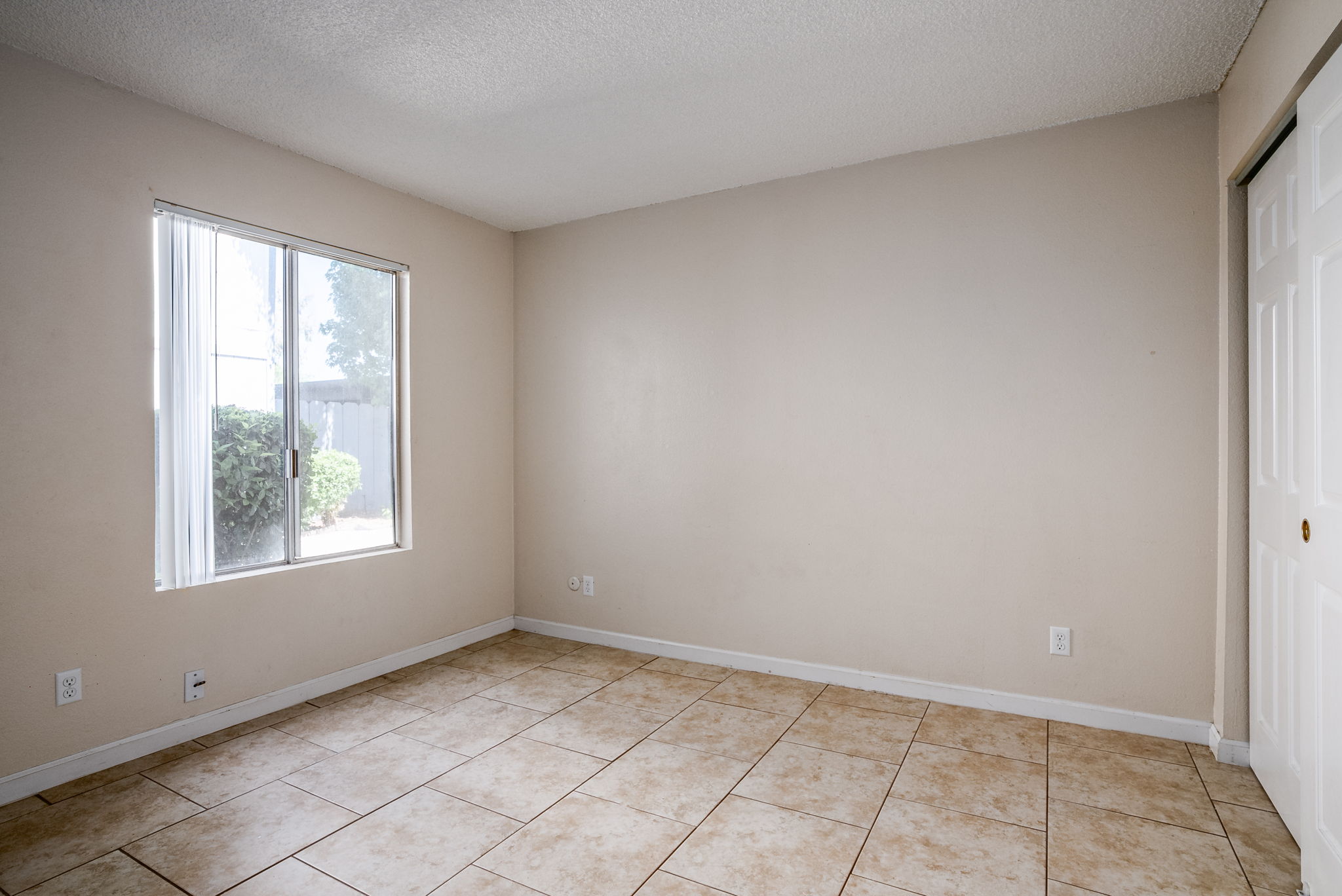 Empty room with beige walls and tiled floor. A window on the left side lets in natural light, offering a view of greenery outside. The room has a single white closet door on the right, creating a simple and open atmosphere suitable for various uses.