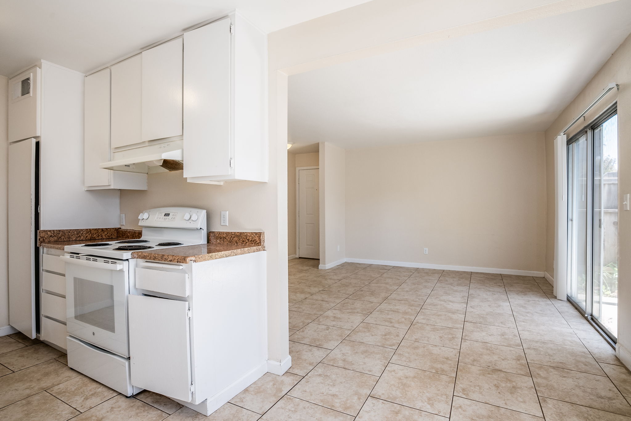 A spacious kitchen featuring white cabinets, a stove, and countertop with a brown speckled pattern. The floor is tiled in a light color. Adjacent to the kitchen is a bright, empty living area with light walls and large sliding doors leading outside, creating an open and airy atmosphere.
