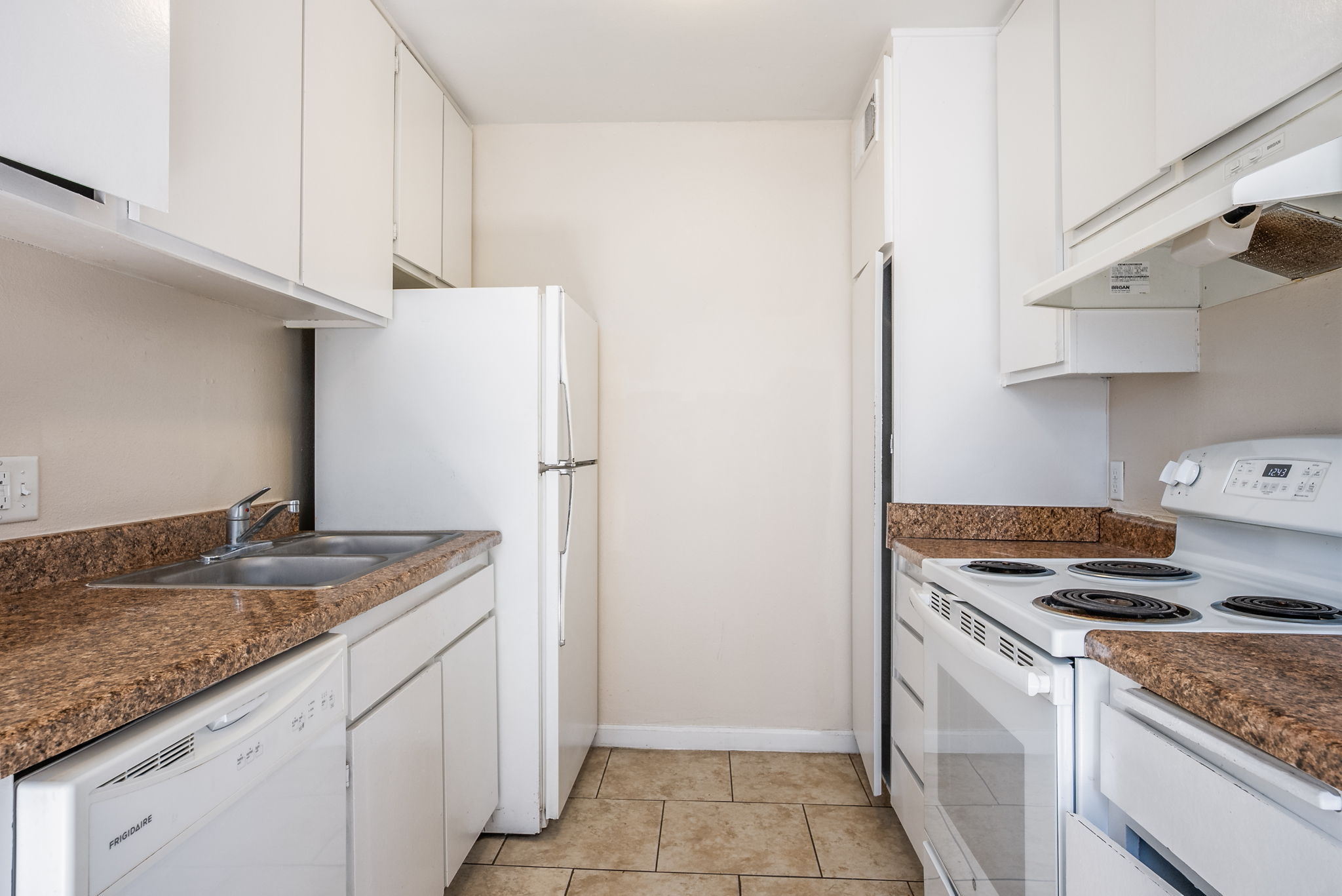 A small, modern kitchen featuring white cabinets, a stainless steel refrigerator, a double-basin sink, a stove with an oven, and a microwave. The countertops are a light brown color, and the walls are painted cream. The layout is efficient, with appliances arranged closely for convenience.