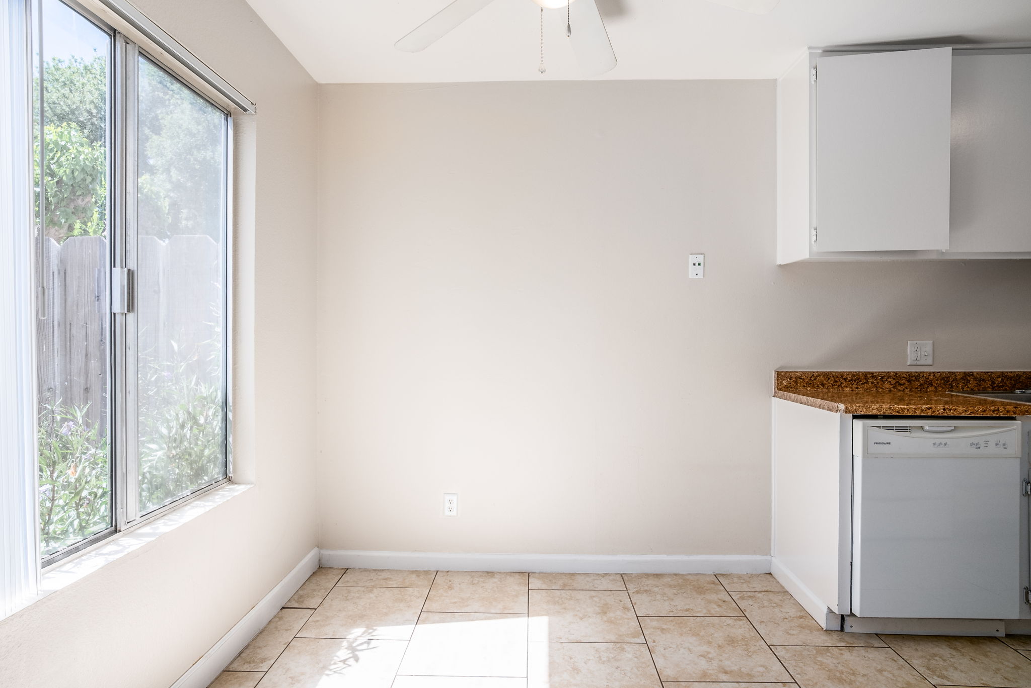 Bright and spacious kitchen interior featuring a large window allowing natural light, beige walls, and tiled flooring. The kitchen includes a white dishwasher next to a granite countertop with cabinets above. The room has a ceiling fan and minimal furnishings, providing an open and airy atmosphere.