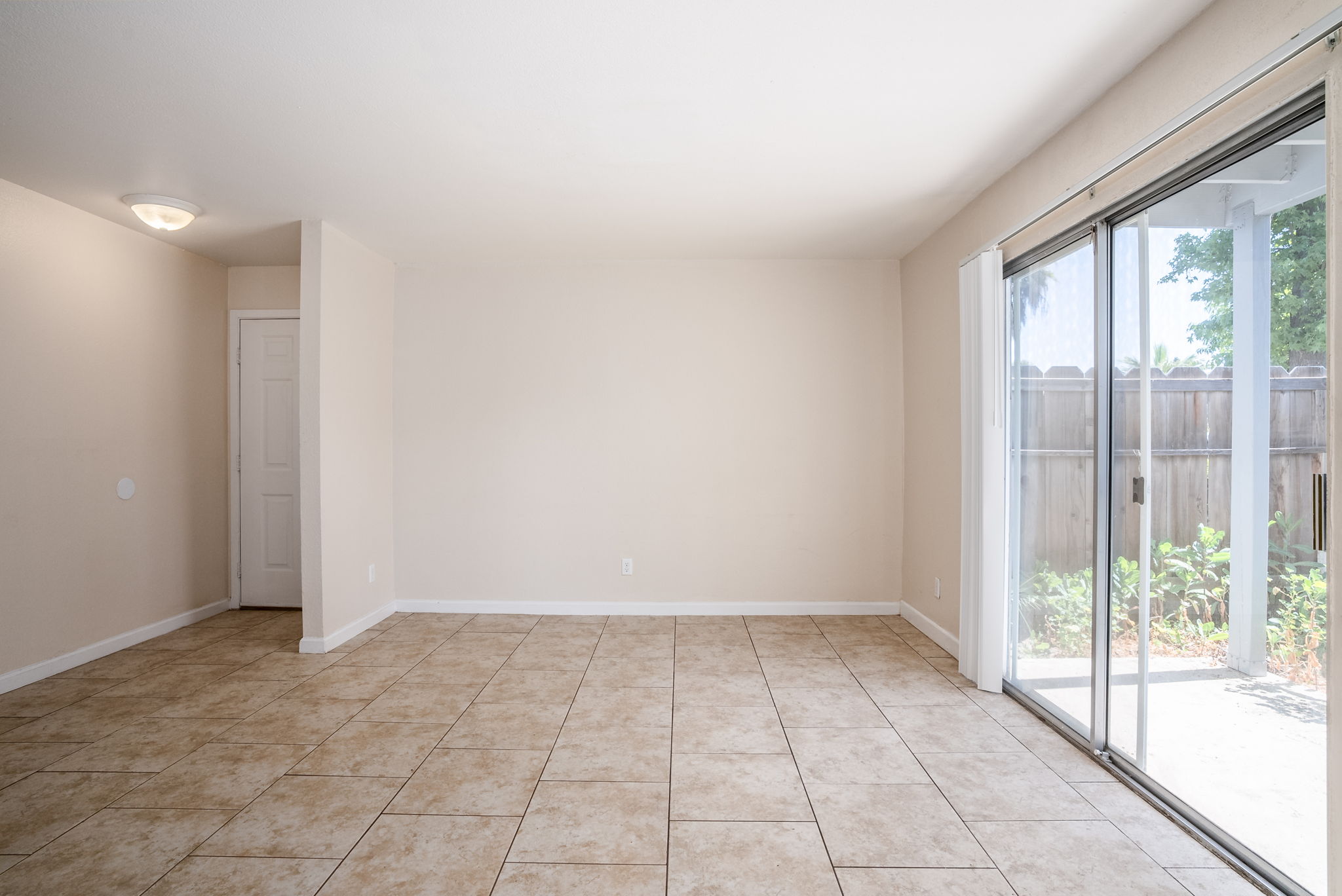 A spacious, empty room with tiled flooring and light-colored walls. It features a large sliding glass door leading to an outdoor area, allowing natural light to enter. There is also a small closet or storage space visible on one side. The overall ambiance is bright and open.