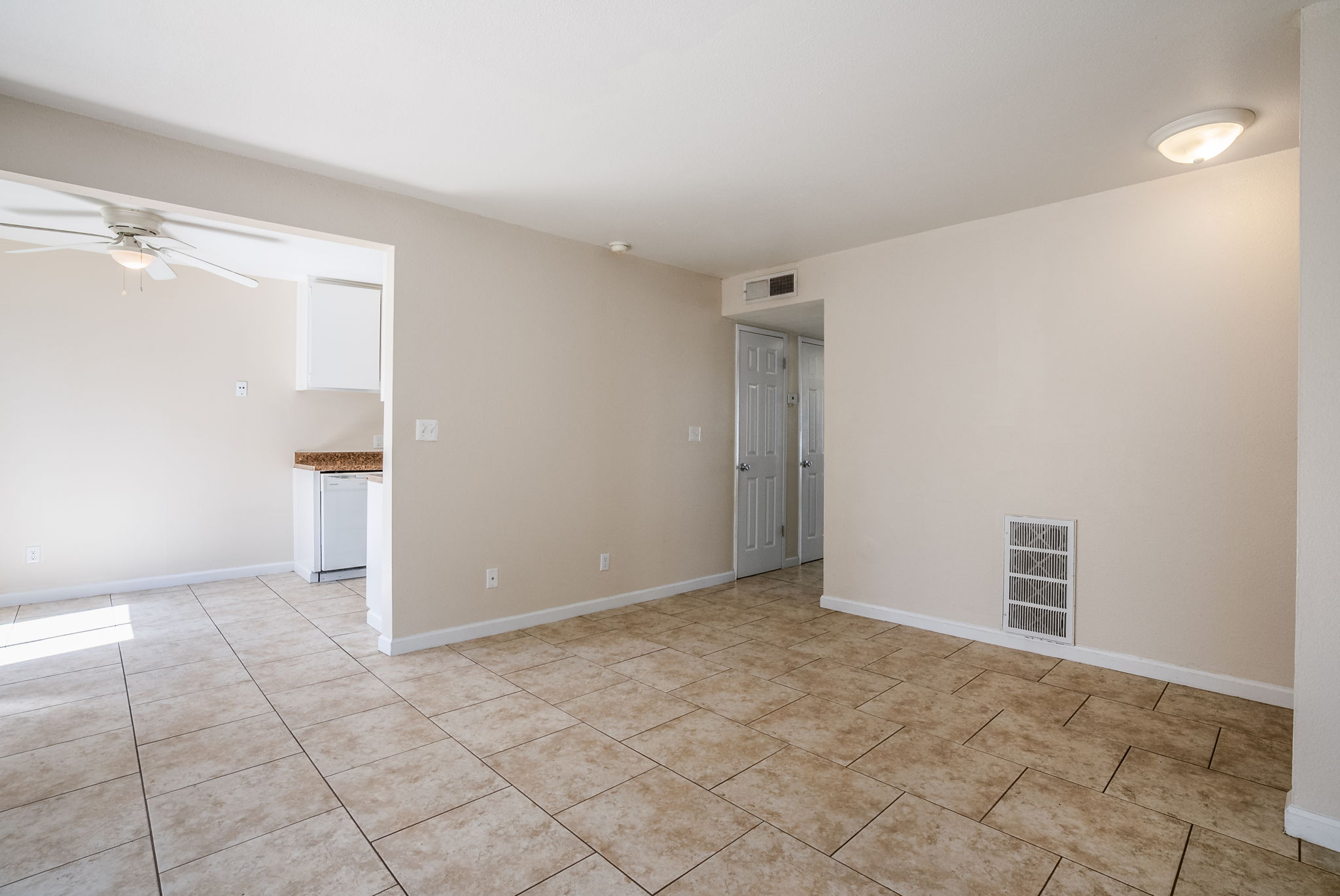 Spacious interior of a light-filled room featuring beige walls, tiled flooring, and a ceiling fan. The open layout includes a doorway leading to another area, with a small kitchen visible in the background. Natural light enters through windows, creating a bright and inviting atmosphere.