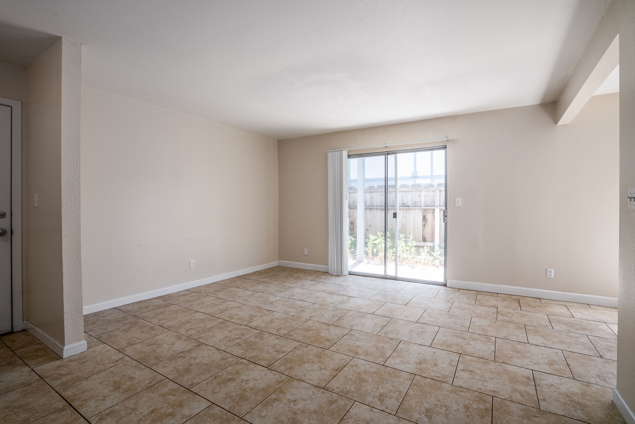 A spacious, unfurnished living room with beige walls and large, tiled floor. A sliding glass door leads to an outdoor area, and natural light fills the space. The room has no furniture and appears clean and minimalistic, ideal for personal customization.