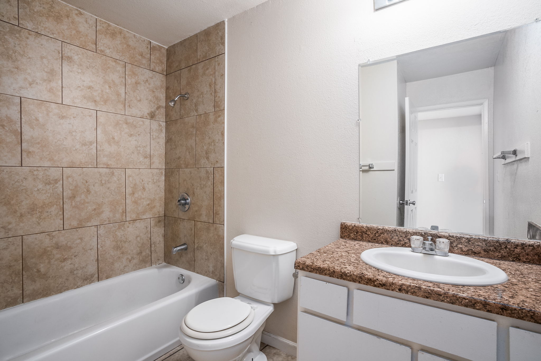 A small bathroom featuring beige tiled walls, a bathtub with a showerhead, a white toilet, and a granite countertop with a sink. A mirror is mounted above the sink, and there is an open door leading to another room. The overall decor is simple and functional.