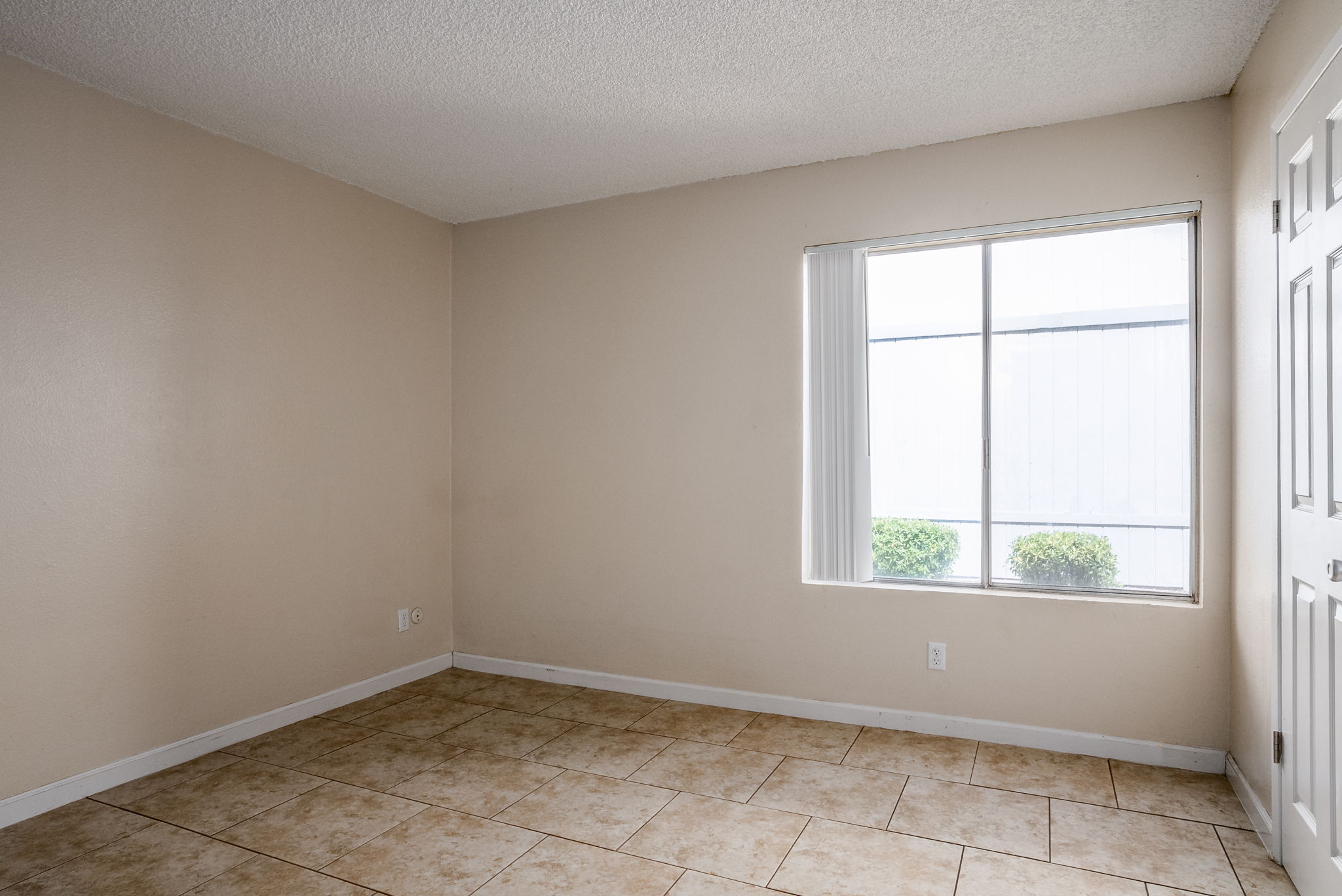 A small, empty room with beige walls and tiled flooring. There is a large window on one wall letting in natural light, along with a door on the adjacent wall. The window features a view of green shrubs outside. The space appears clean and ready for potential furnishings or decoration.