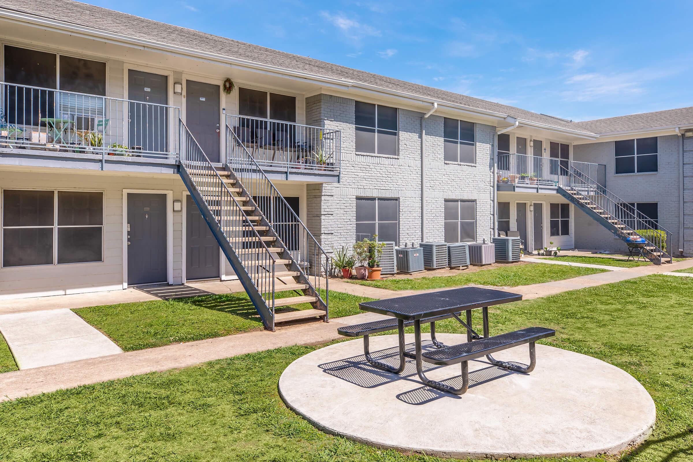 A modern apartment complex with two levels, featuring multiple entrances, stairs leading to the upper floor, and a central grassy area. In the foreground, there's a round picnic table on a concrete slab, surrounded by well-maintained landscaping and air conditioning units. Clear blue sky overhead.