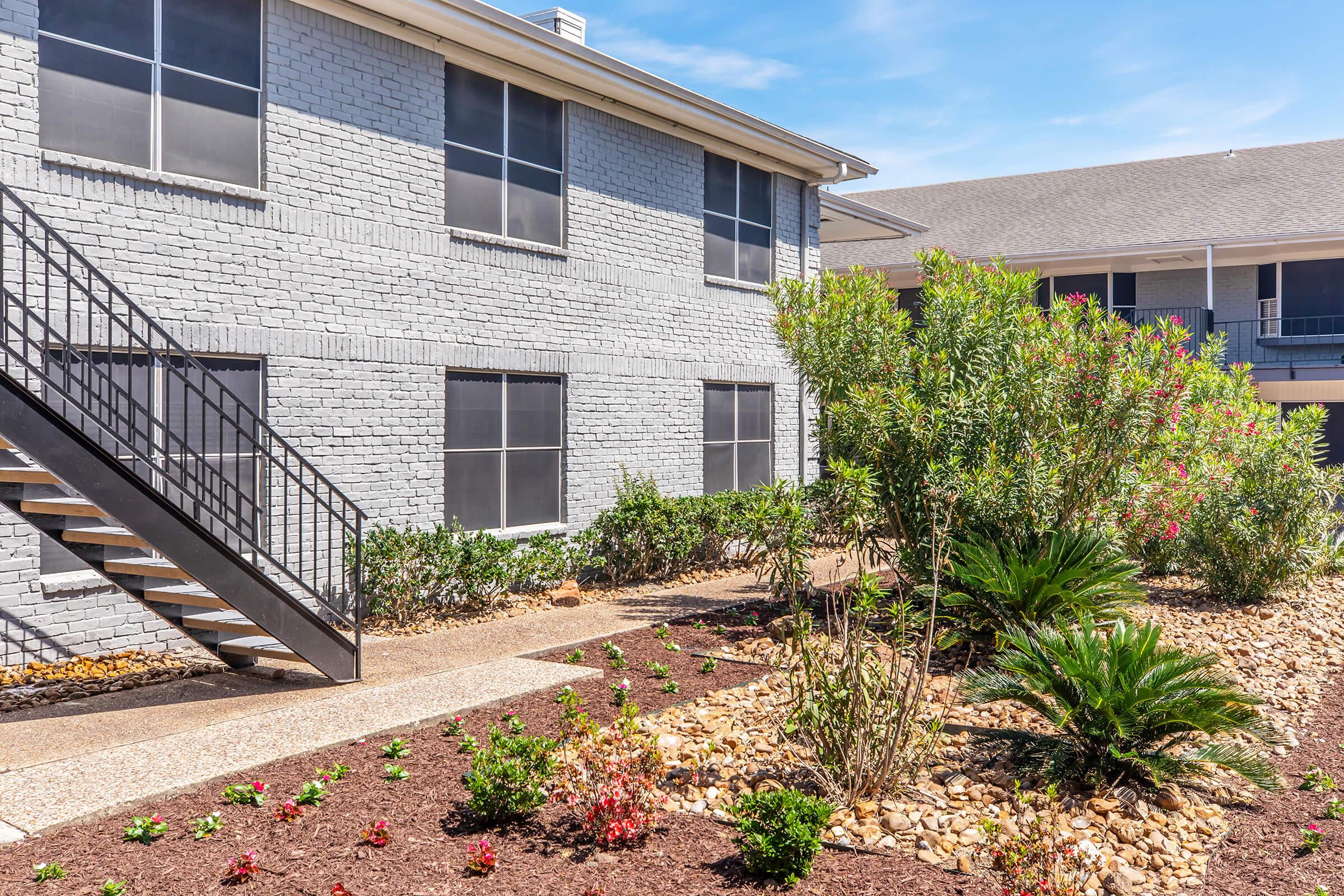 A view of a two-story apartment building with gray brick walls and multiple windows. A decorative garden features various plants, shrubs, and a pebble walkway. An outdoor staircase leads to the second floor, set against a clear blue sky.