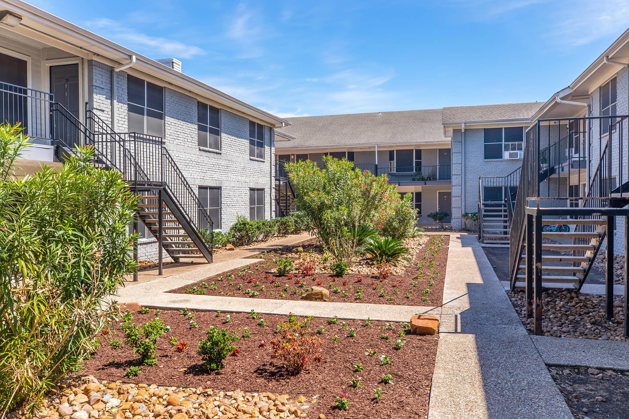 A well-maintained courtyard of an apartment complex featuring freshly landscaped flower beds with plants and mulch. There are two sets of stairs on either side leading to the upper floors of the buildings. Bright blue sky is visible overhead, enhancing the inviting atmosphere of the outdoor space.
