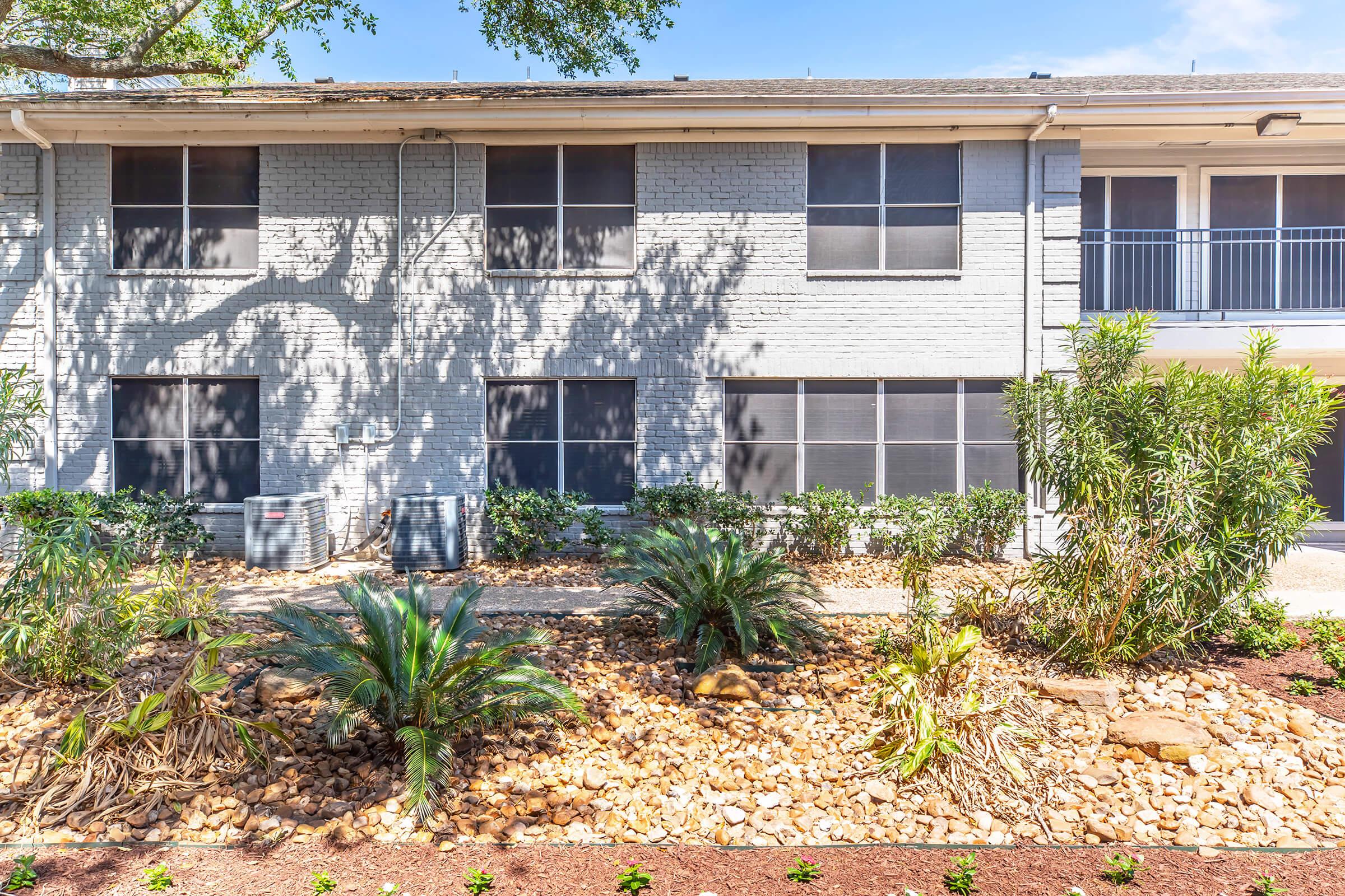 Exterior view of a residential building with a light gray brick facade. The image features several windows and an entrance, surrounded by landscaping including palms and rocks. Sunlight casts shadows from nearby trees, creating a bright and inviting atmosphere.