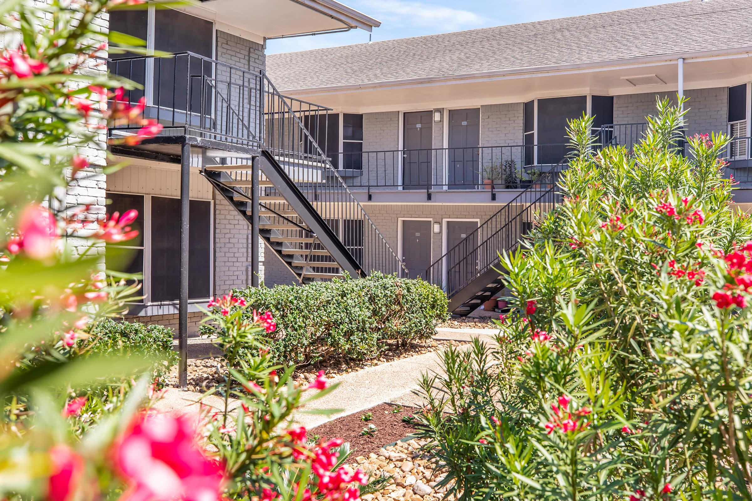 A view of a multi-story apartment complex featuring a stairway and balcony. Lush green shrubs and vibrant pink flowers are in the foreground, enhancing the outdoor space. The building has gray brick exteriors and multiple entrances, with a clear blue sky in the background.