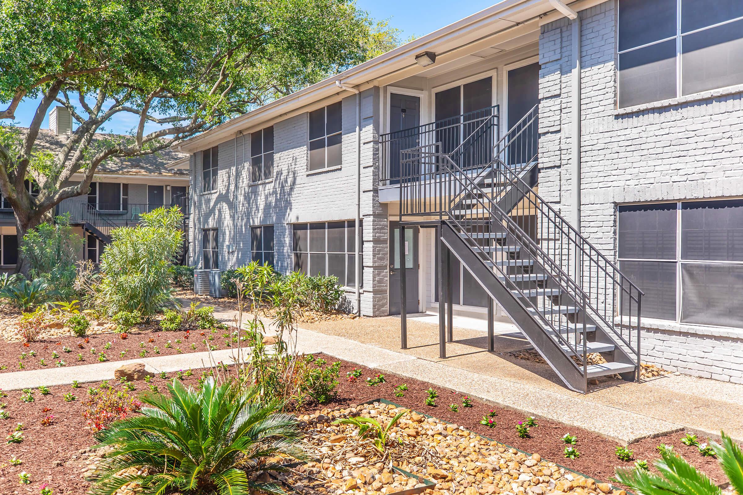 A modern, two-story apartment building with a gray exterior and a staircase leading to the second floor. Lush landscaping, including various plants and flowers, surrounds the building, with a pathway made of gravel leading to the entrance. Bright sunlight illuminates the scene.