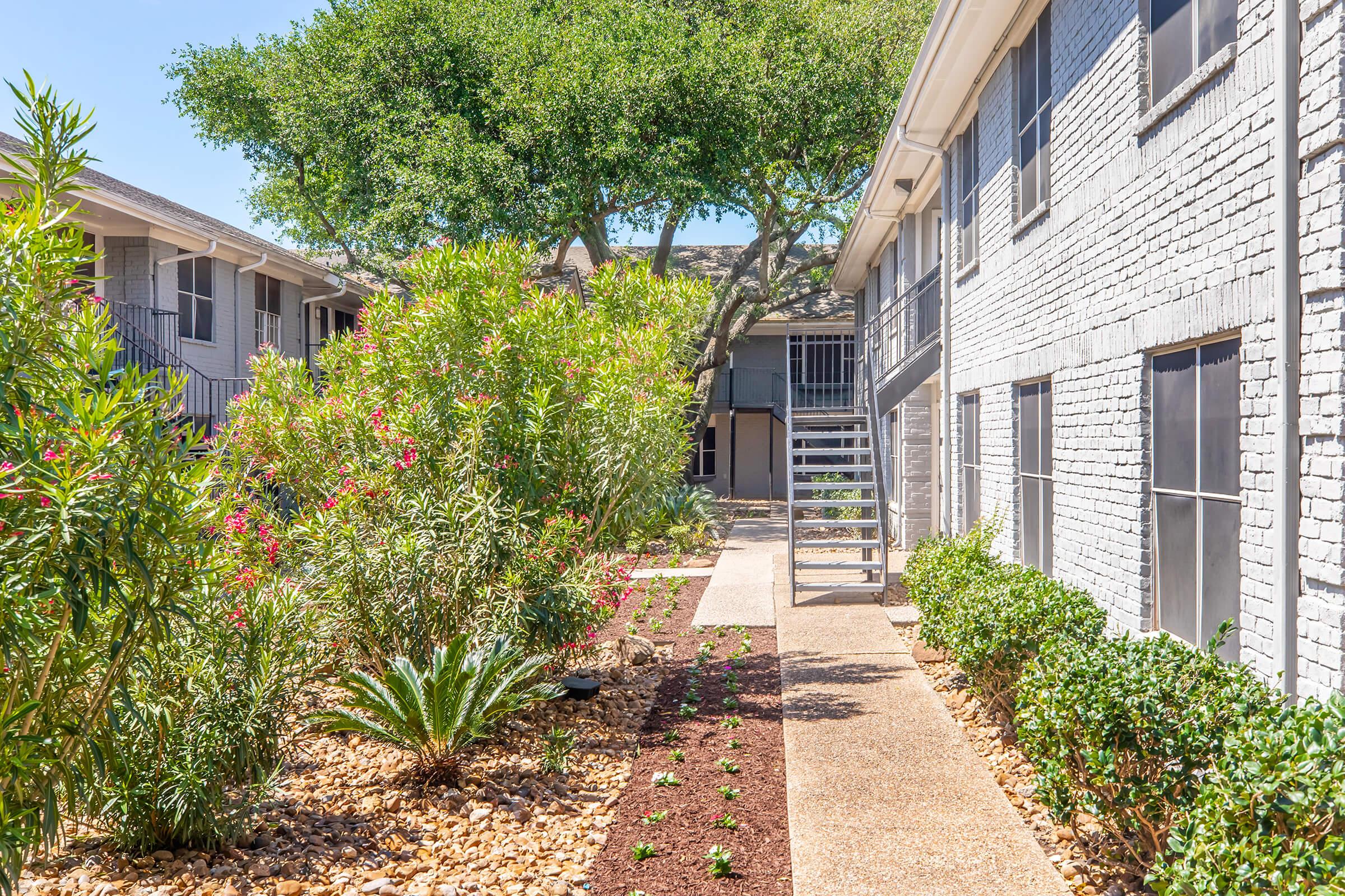Lush courtyard of an apartment complex featuring green plants, flowering bushes, and a pebble pathway. In the background, there are two multi-story buildings with balconies, and a staircase is visible on the right side. Bright, sunny day contributing to a vibrant and welcoming atmosphere.