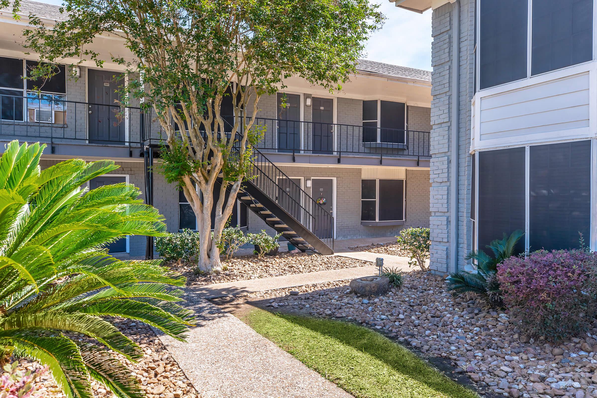A landscaped courtyard featuring palm trees, shrubs, and a stone path leading to an apartment complex. There are two buildings with balconies, greenery, and a staircase, creating a welcoming outdoor space in a residential setting. Bright, sunny skies add to the inviting atmosphere.