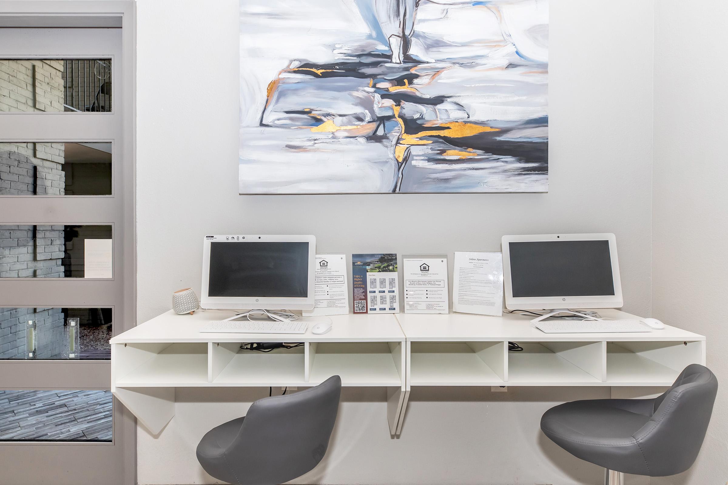A modern workspace featuring two desktop computers on a white table with storage compartments. Accompanying the computers are keyboards and a speaker. A large abstract painting in shades of gray and orange hangs above the table. Two gray chairs are positioned at the table, and a glass door leads to another area.