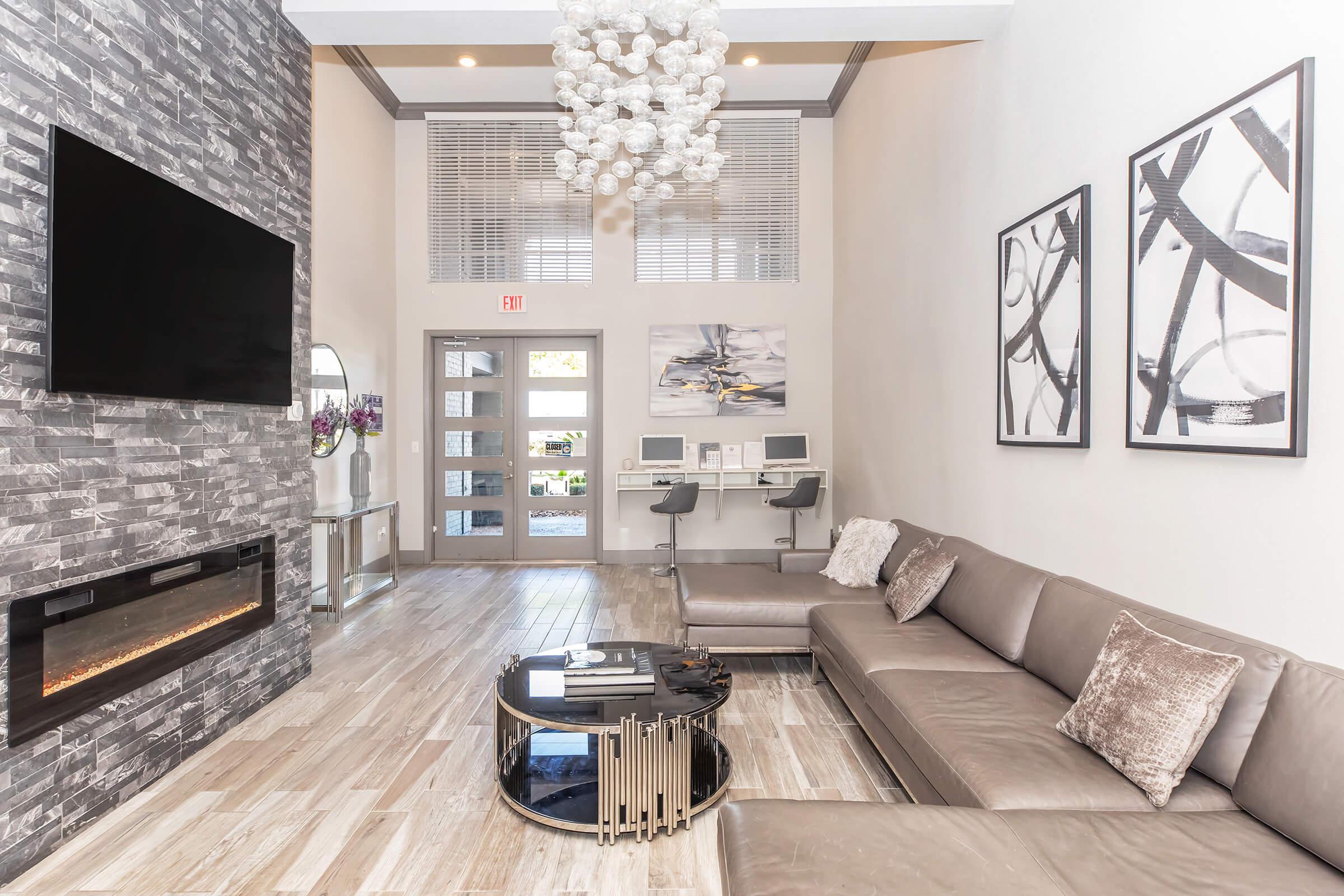 Modern living room with a stone accent wall and a large flat-screen TV. Features a contemporary gray sofa and a sleek coffee table. A striking chandelier hangs overhead, and artwork adorns the walls. There's a work area with computers visible in the corner, contributing to a stylish and functional space.