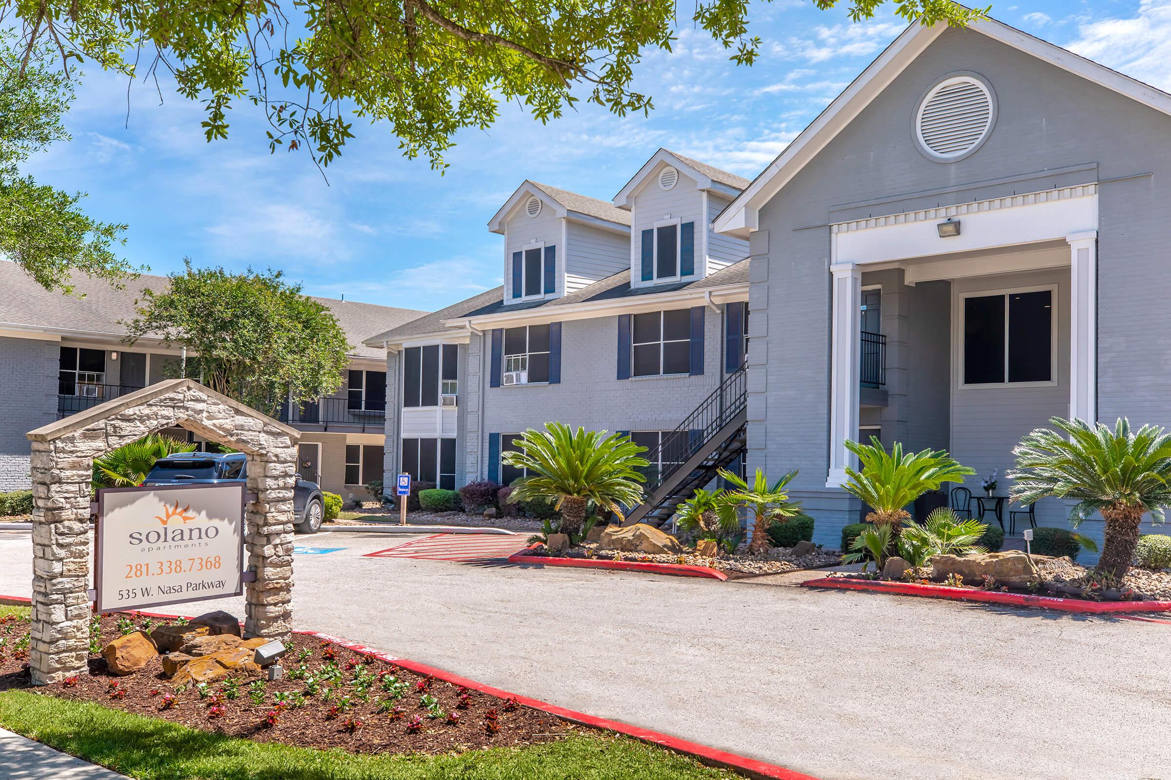 Exterior view of Solano Apartments featuring a modern building design with multiple units. Lush palm trees and landscaping adorn the entrance along with a well-maintained driveway. A sign displaying the name and contact details for the apartments is prominently displayed at the entrance. Clear blue skies add to the bright atmosphere.