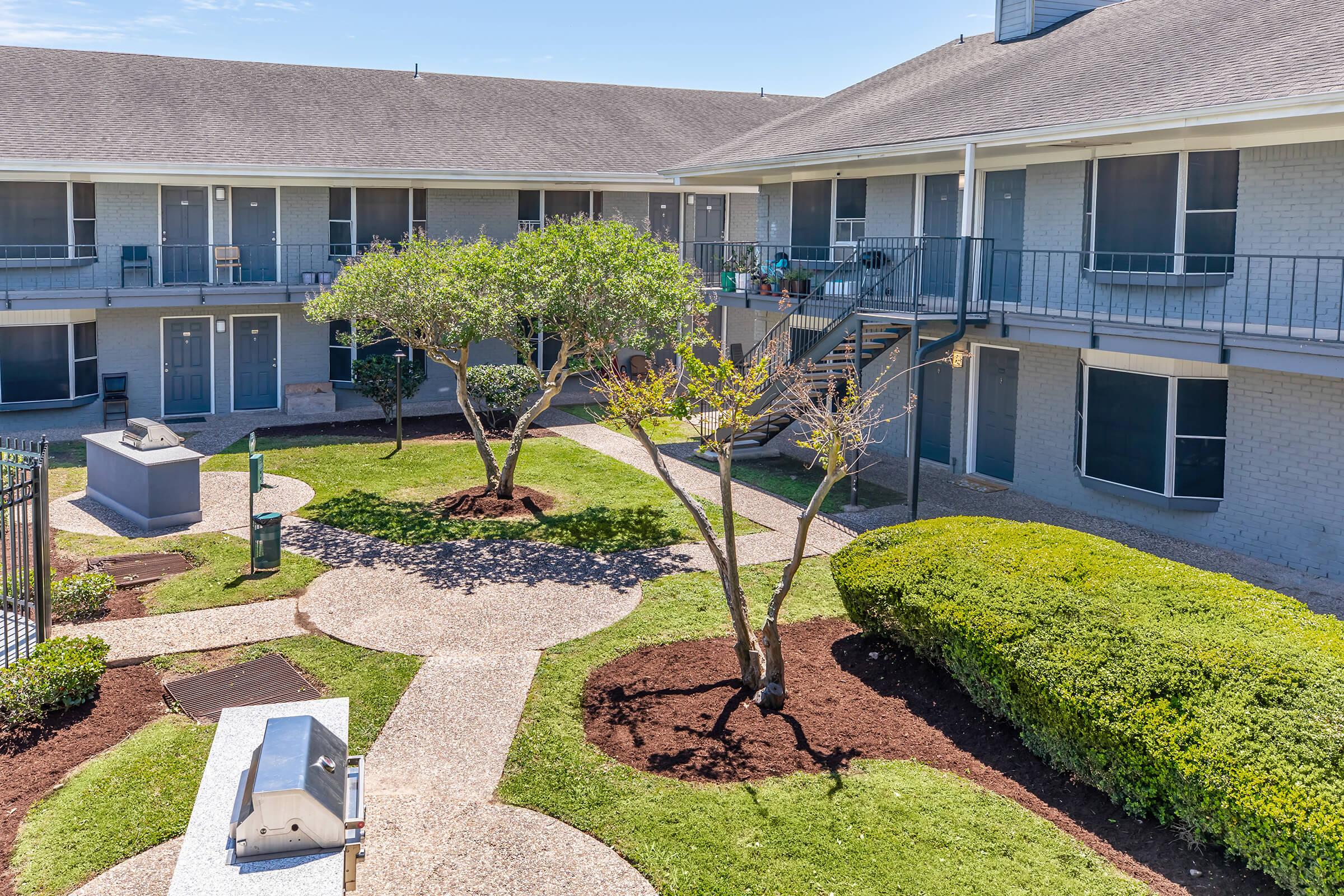 A peaceful courtyard of a residential building featuring neatly trimmed grass, two small trees, and pathways winding through the garden. Several ground-level apartments with windows overlook the area, and some outdoor seating is visible. Bright, sunny weather enhances the inviting atmosphere.