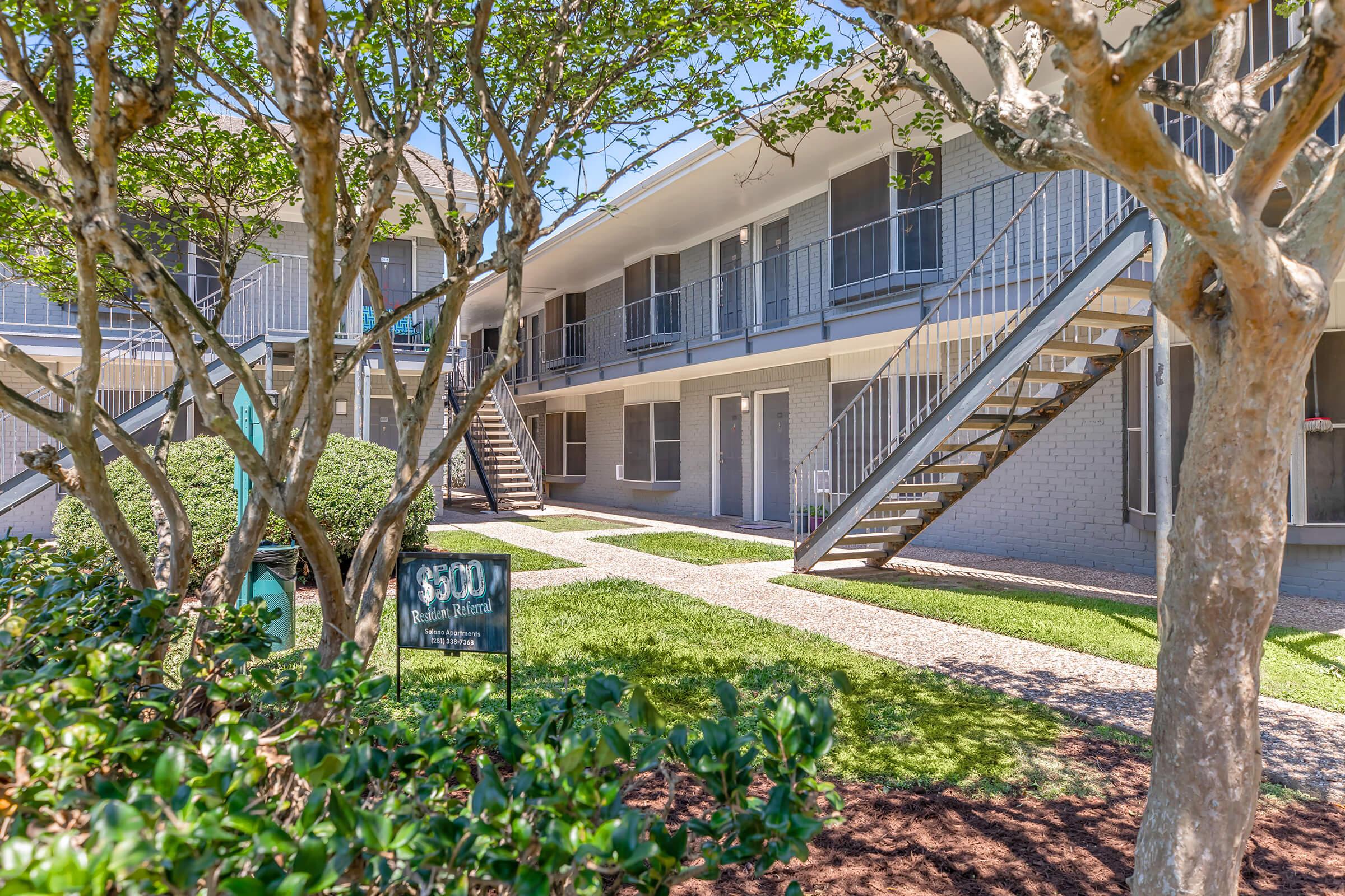 A view of a multi-unit apartment building with two levels, featuring external staircases and well-maintained landscaping. There are small trees and shrubs in the foreground, and a sign indicating a rental price of $500. The sky is clear and blue, creating a bright and inviting atmosphere.