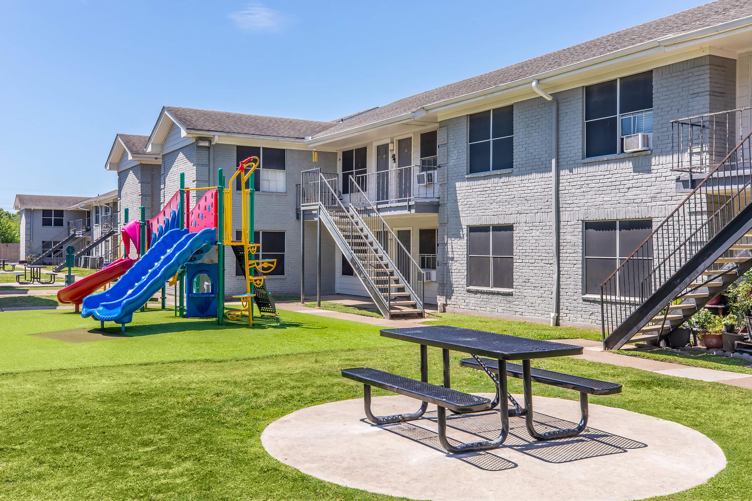 Colorful playground equipment featuring slides and climbing structures in a grassy area, situated in front of two-story apartment buildings. A picnic table is placed on a paved circle nearby, with air conditioning units visible on the building's exterior. Clear blue sky enhances the bright atmosphere.