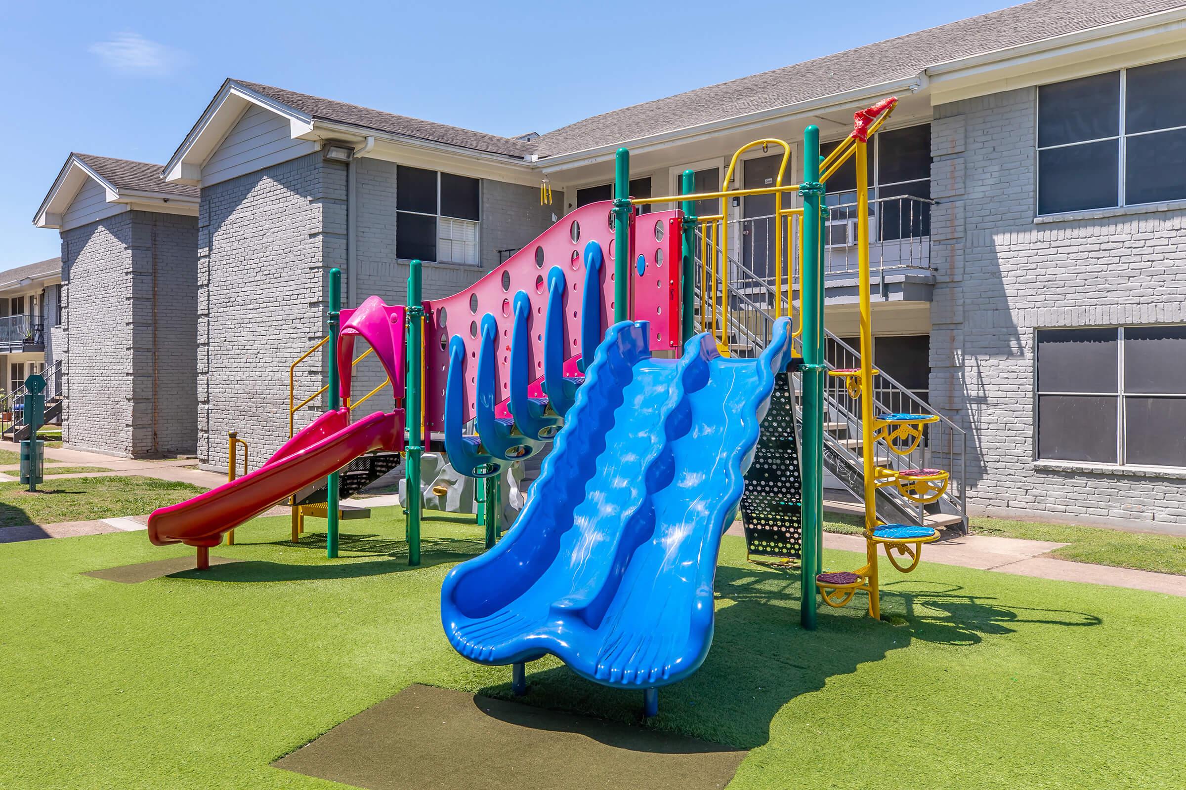 Colorful playground with multiple slides, including a pink and a blue slide, set in a grassy area near residential buildings. The playground equipment features climbing structures and safety railings, inviting children to play and explore. Clear blue sky above enhances the cheerful atmosphere.