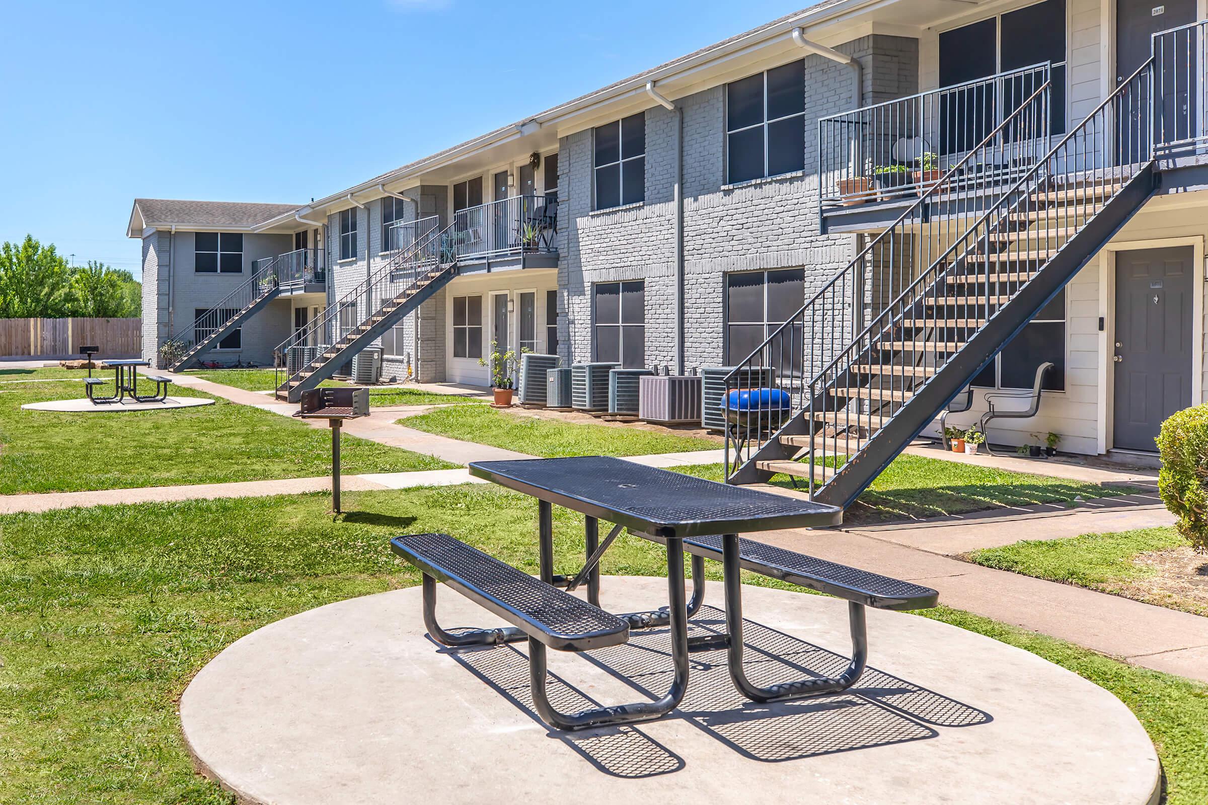 A landscaped courtyard featuring a picnic table with benches on a concrete slab, surrounded by multiple two-story apartment buildings. Staircases lead to upper units, and air conditioning units are visible near the buildings. The sky is clear and blue, creating a pleasant outdoor atmosphere.