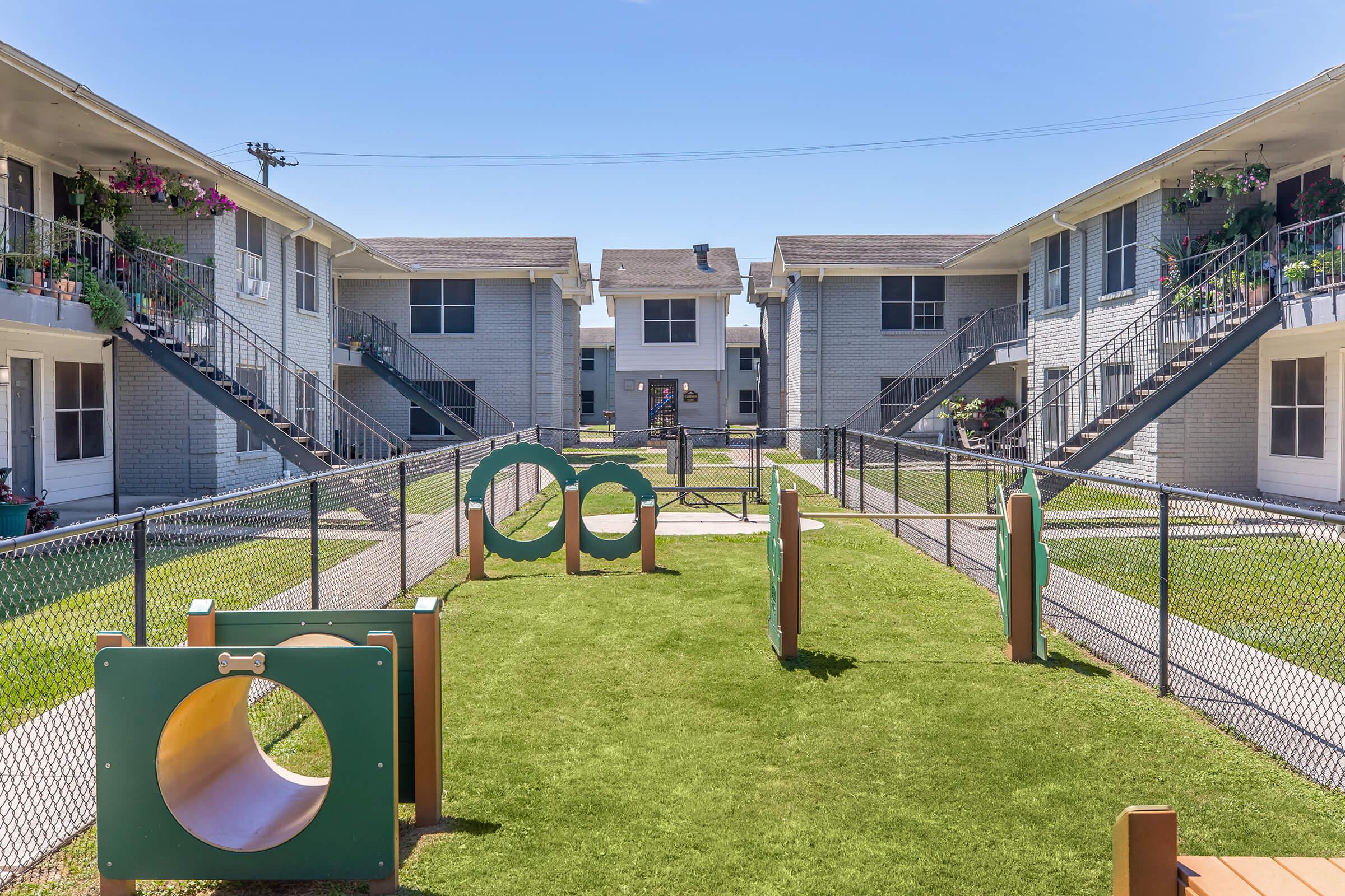 A view of an outdoor area featuring green grass and playground equipment, including circular tunnels and climbing structures, surrounded by a fence. In the background, two multi-story apartment buildings with balconies are visible under a clear blue sky.