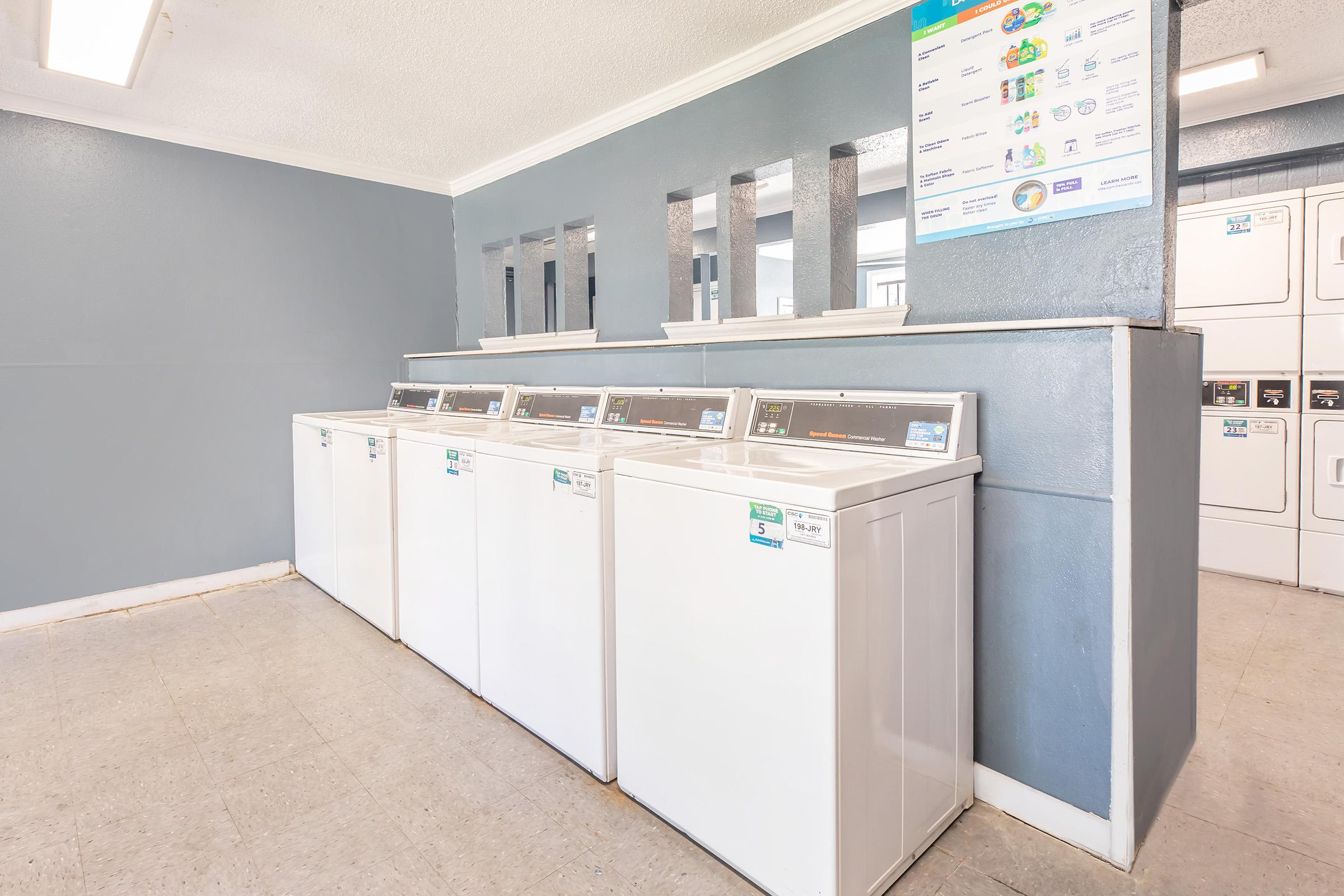 A clean and well-lit laundry room featuring four white washing machines side by side against a gray wall. Mirrors are placed above the machines, and a bulletin board with laundry instructions and tips is displayed on the wall. The flooring is light-colored and the space appears organized and inviting.