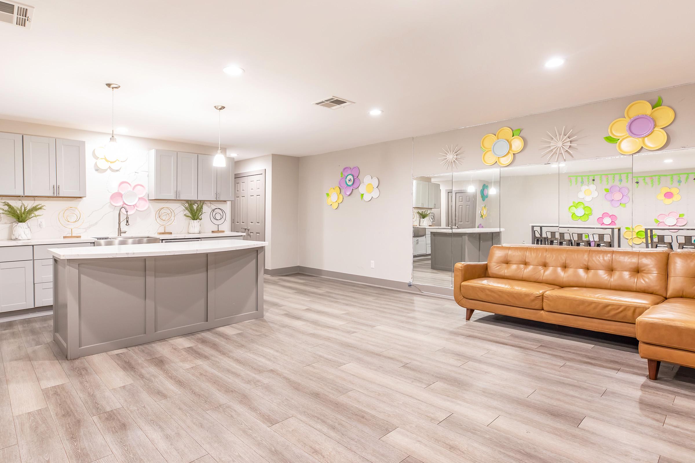 A modern, spacious interior of a kitchen and living area featuring light gray cabinets, a large island with seating, and a tan leather sofa. Colorful flower decorations adorn the walls, adding a cheerful touch to the room, which has wood-like flooring and bright lighting.