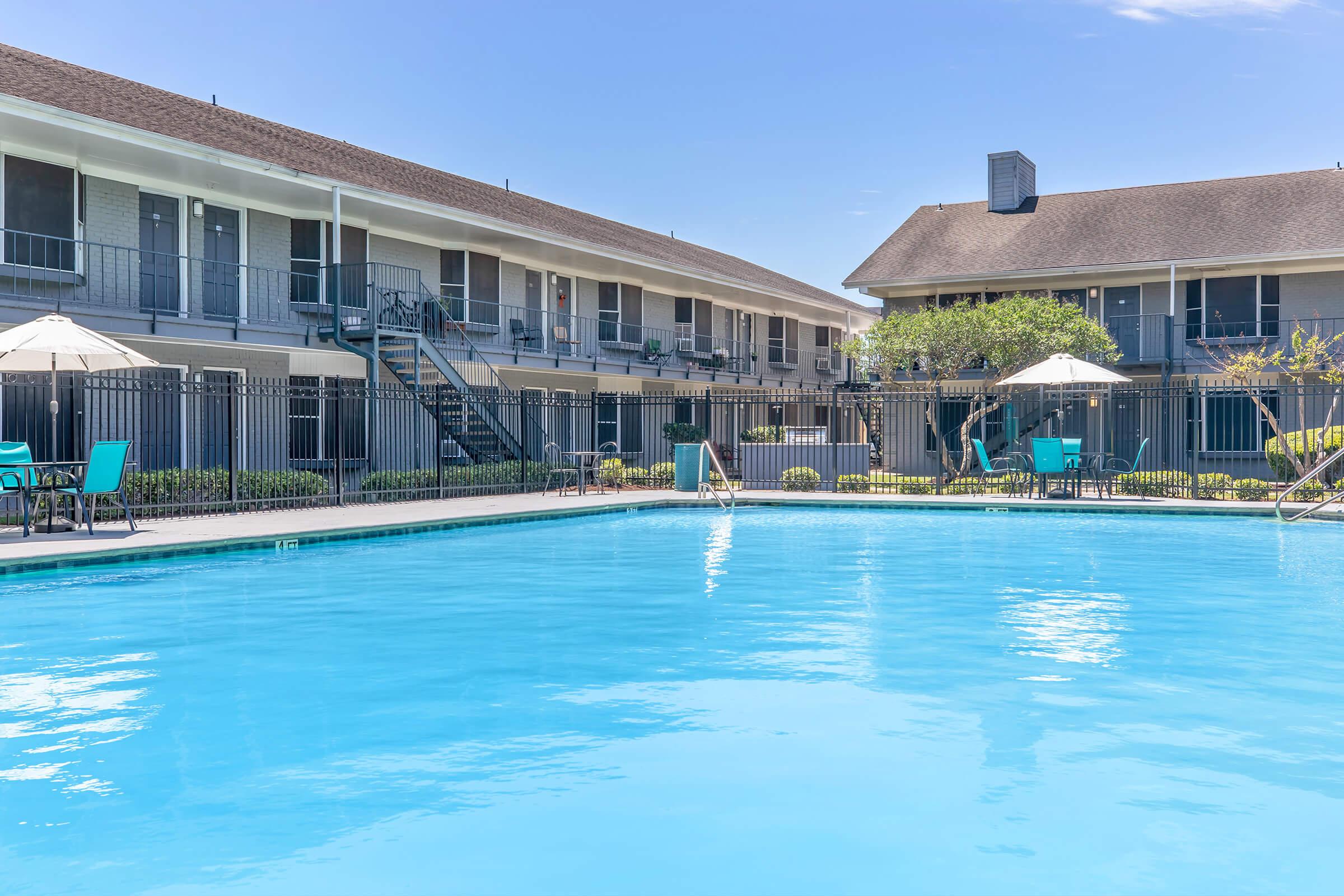 A clear blue swimming pool surrounded by a fence, with lounge chairs and umbrellas. In the background, there are two multi-story buildings with balconies overlooking the pool area. The sky is bright and sunny, enhancing the inviting atmosphere of the scene.