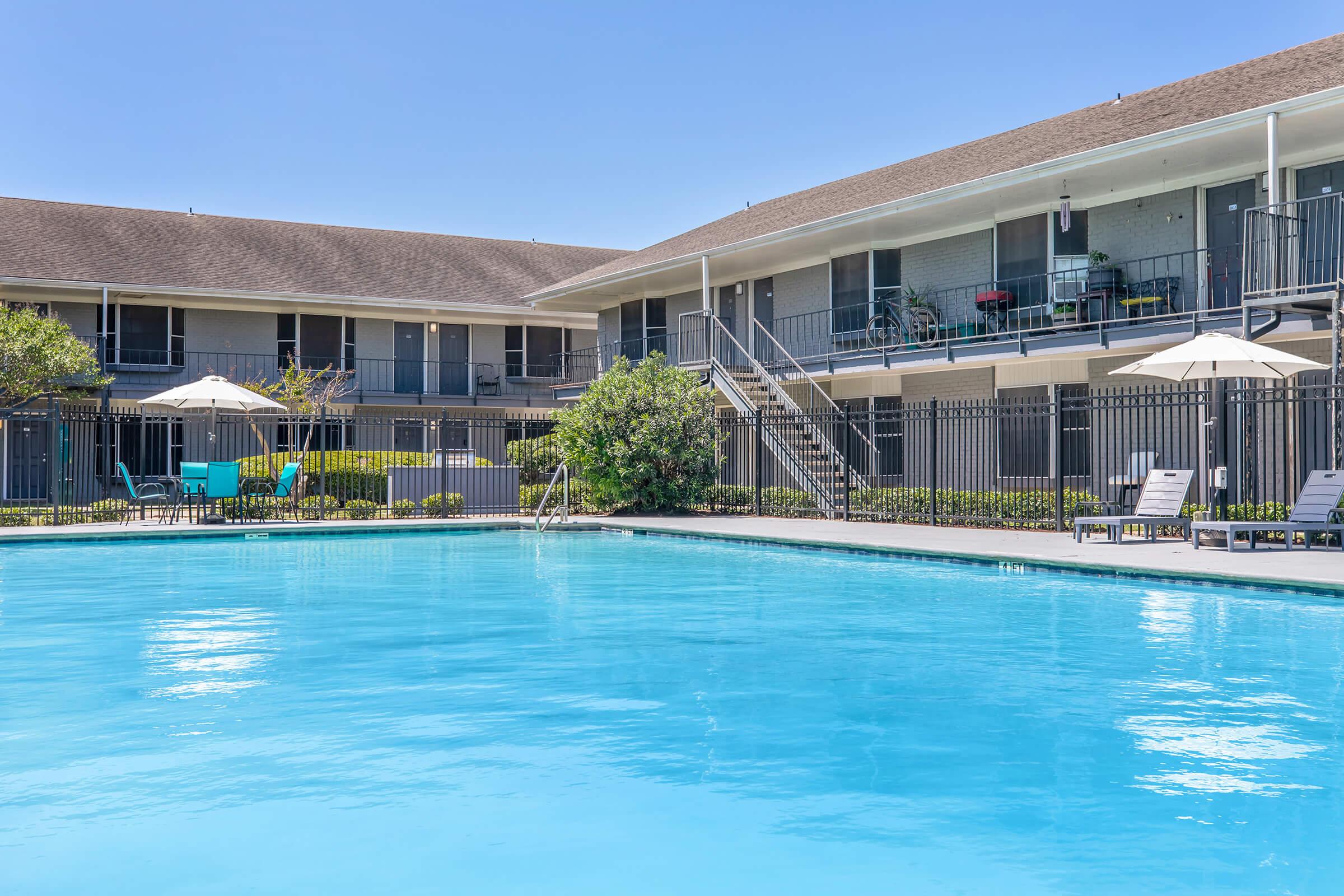 A clear blue swimming pool surrounded by lounge chairs and umbrellas, situated in a courtyard between two multi-story buildings. The buildings have balconies and a well-maintained garden area, with sunny skies overhead.