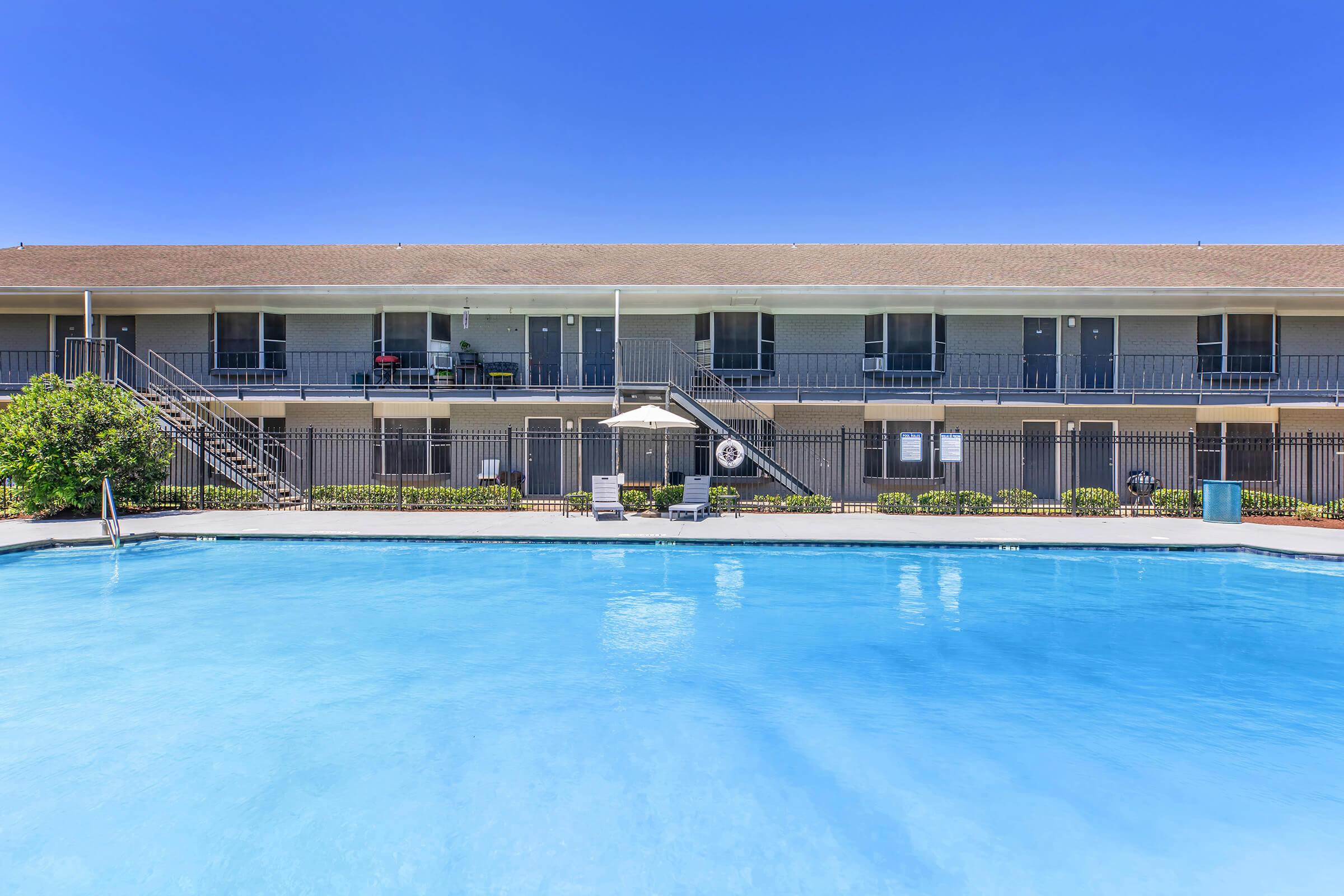 A clear blue swimming pool in front of a two-story building with balconies. The building features several doors and outdoor seating areas. Lush green landscaping surrounds the pool area, and a large umbrella provides shade near the water's edge, all under a bright blue sky.