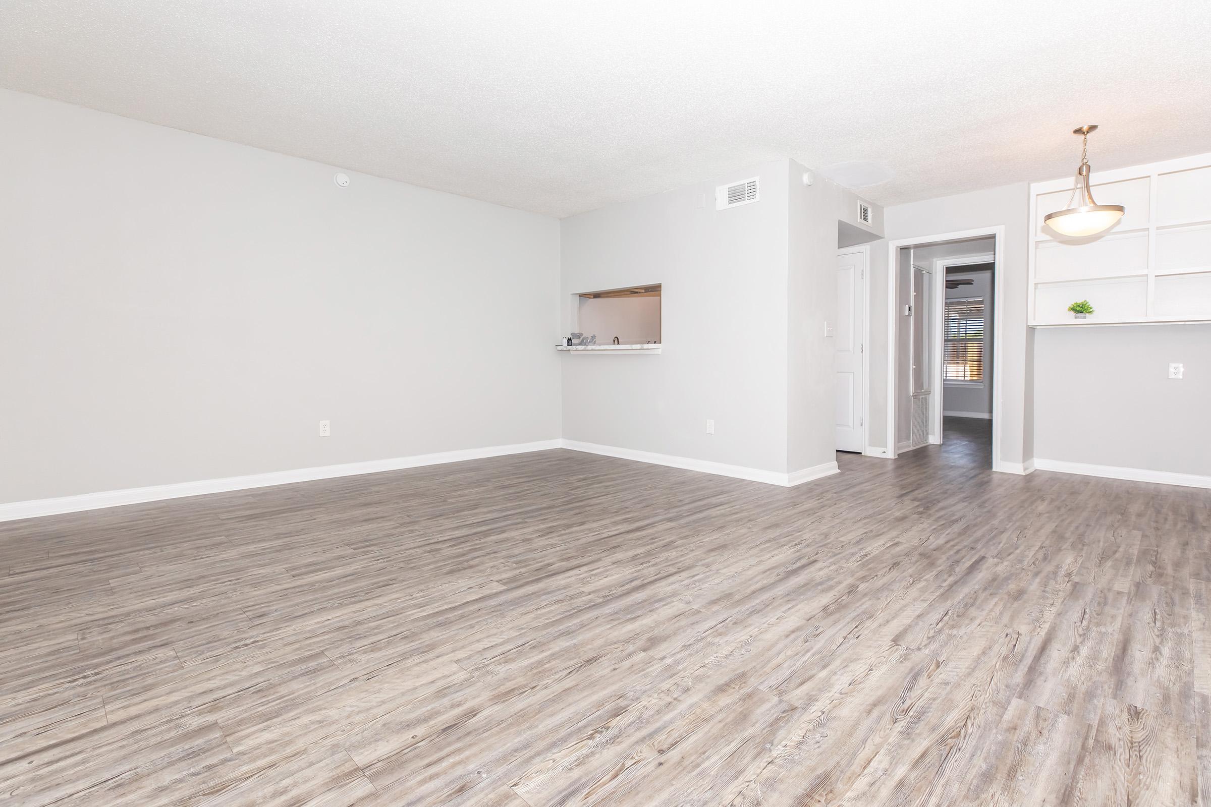 Spacious and modern living room with light gray walls and wood-like flooring. Features an open layout with a small kitchen area visible in the background. Natural light illuminates the space, highlighting the clean design. There's a doorway leading to another room and a pendant light fixture.
