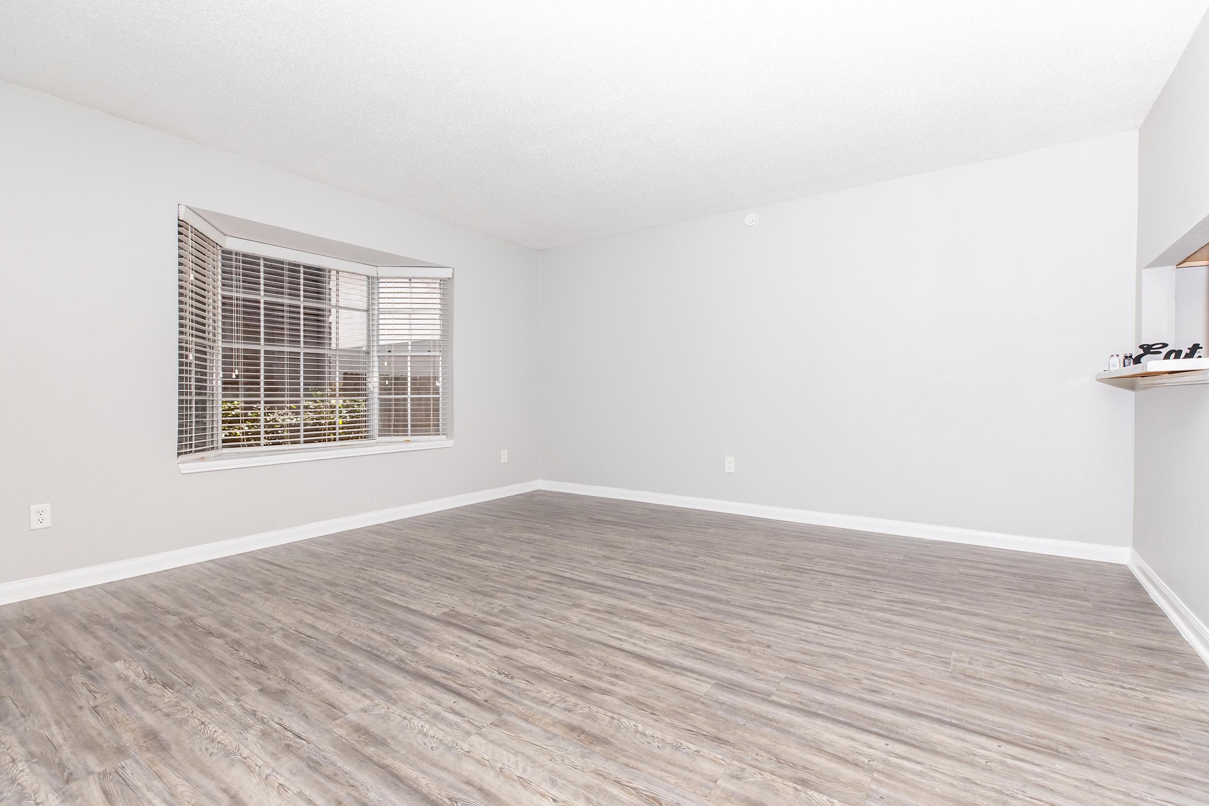 A spacious, empty room with light gray walls and wooden laminate flooring. It features a large, multi-pane window with white blinds allowing natural light in. There is a small opening on one side that leads to another space, possibly a kitchen or dining area. The overall atmosphere is bright and inviting.