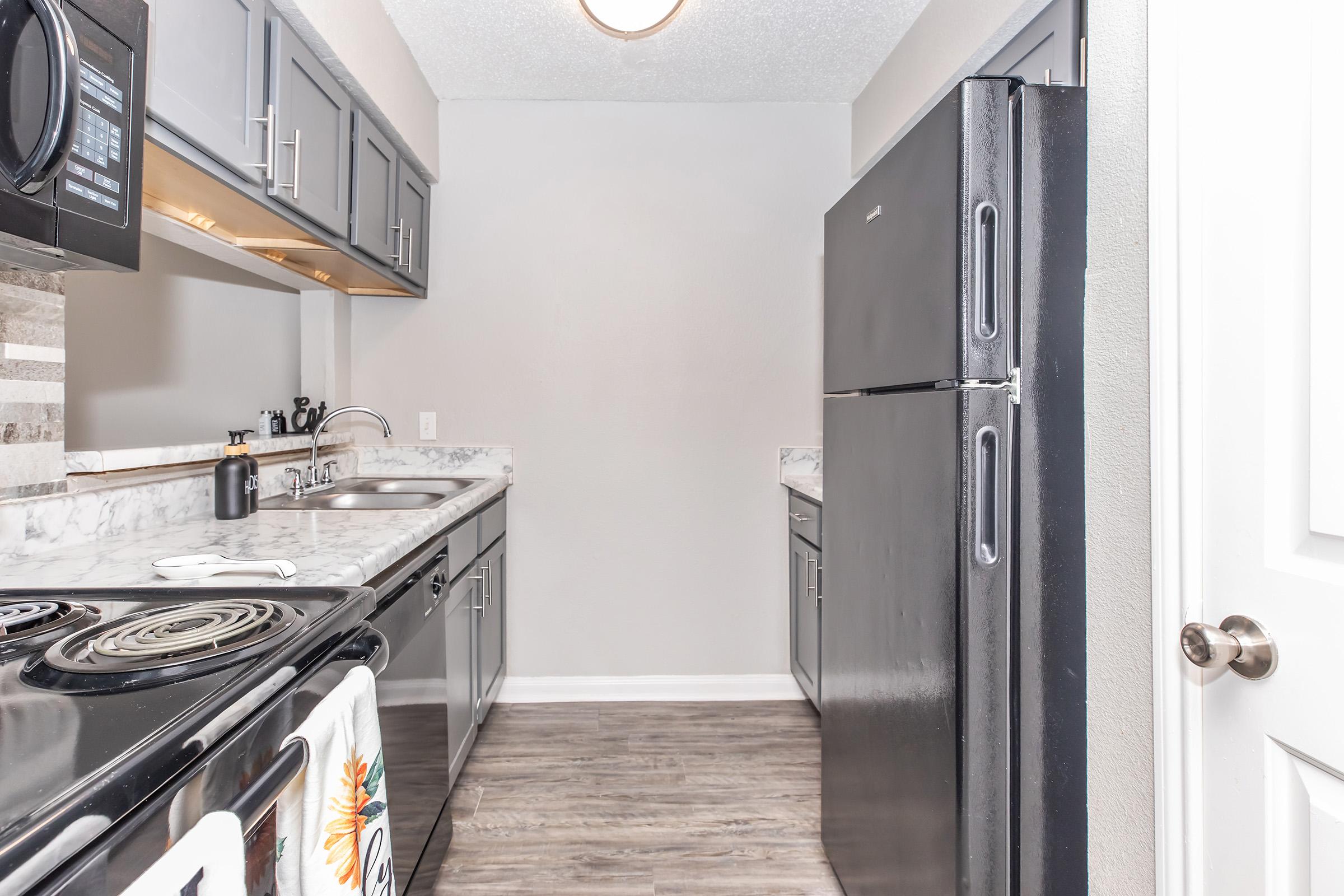 A modern kitchen featuring gray cabinets, a black refrigerator, a stove with an oven, and a double sink. The countertops are white with gray veining, and there's a dish towel hanging on the stove. The walls are painted in a light color, adding to the bright, open feel of the space.