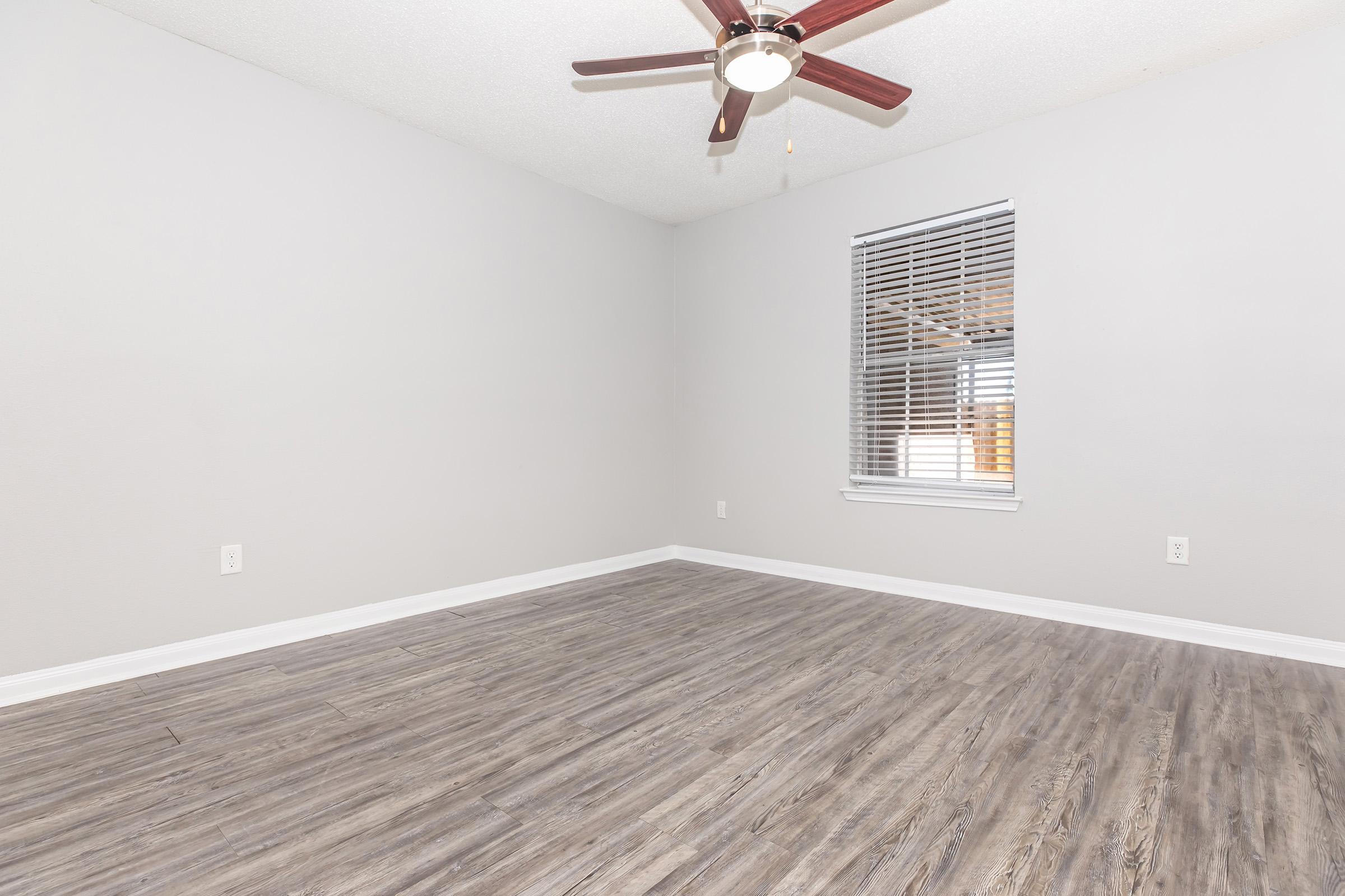 A spacious room with light gray walls and a ceiling fan. It features a wooden floor with a natural finish and a window with horizontal blinds, allowing natural light to enter. The room is empty, showcasing its clean and modern design.