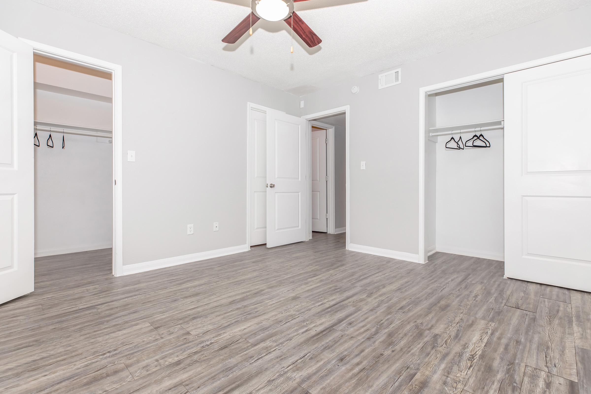 A spacious, empty interior room featuring a ceiling fan, light gray walls, and wooden laminate flooring. Two open doors lead to closets, one with hangers. The room has a bright and airy feel, illuminated by natural light.