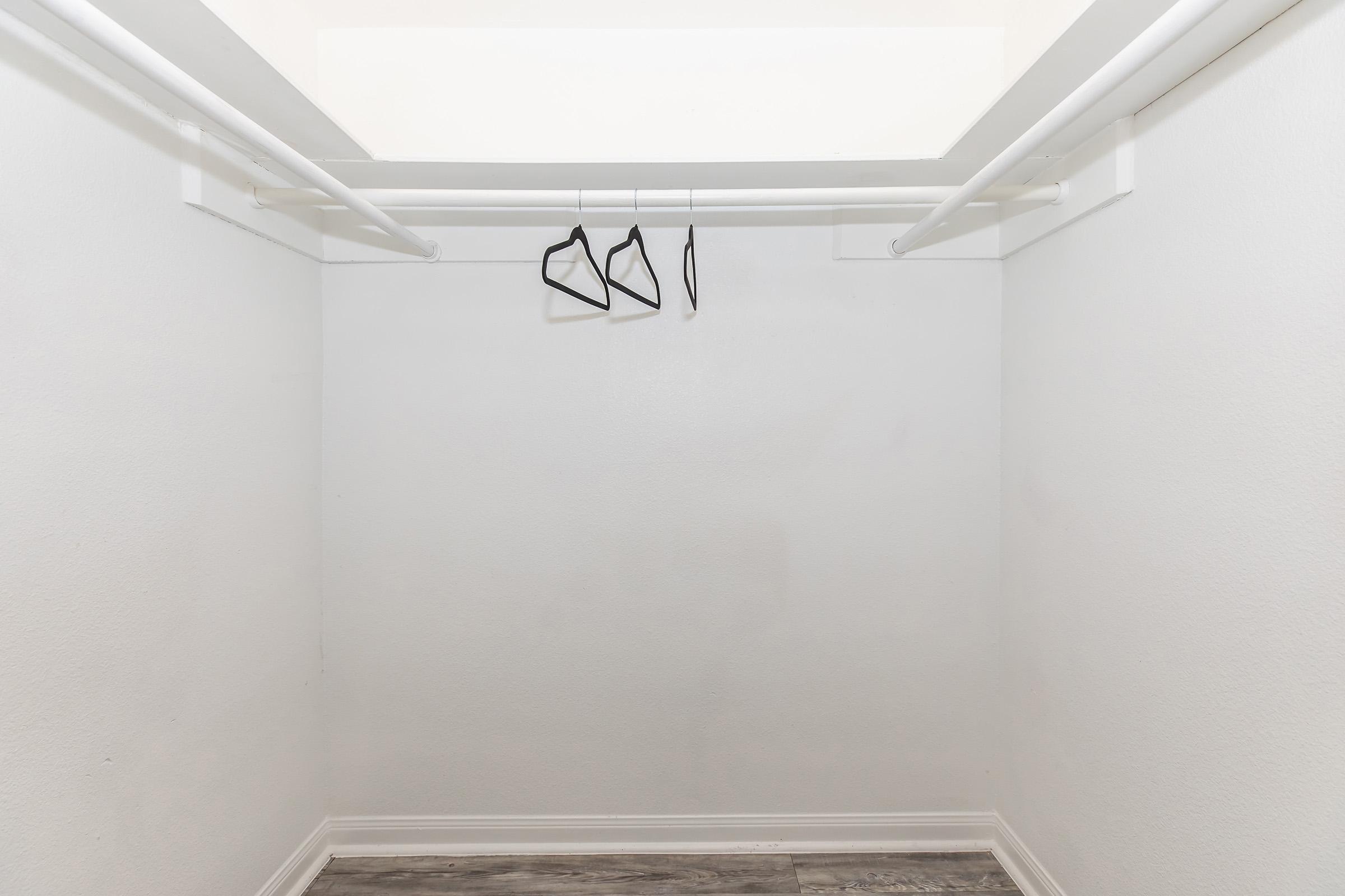 A small, empty closet with two hanging rods. Three black hangers are suspended from the upper rod. The walls are plain and white, and the floor has a light-colored finish. The space appears tidy and uncluttered.