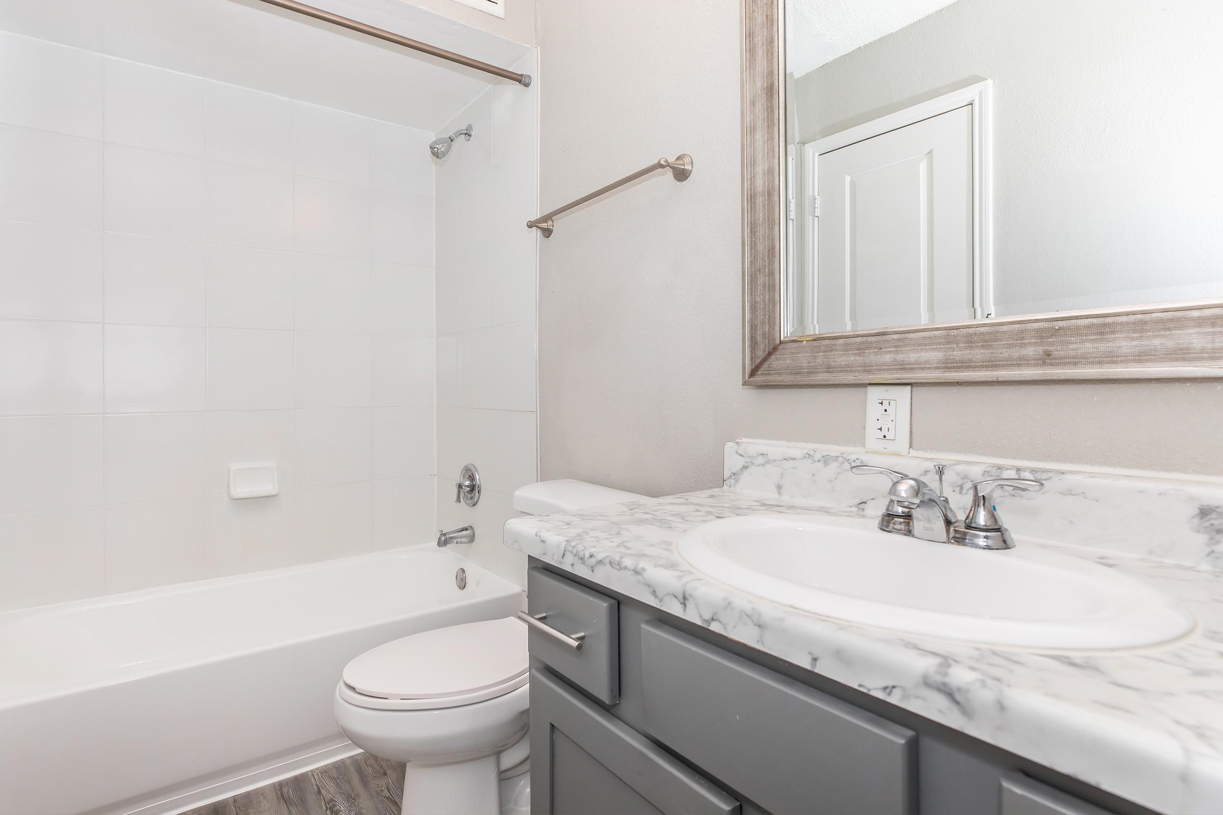 A modern bathroom featuring a white tub and shower, a marble countertop with dual sinks, and a decorative mirror. The walls are painted in a light color, and there is a towel bar mounted on the wall. The floor is styled with wood-like tiles, creating a clean and inviting space.