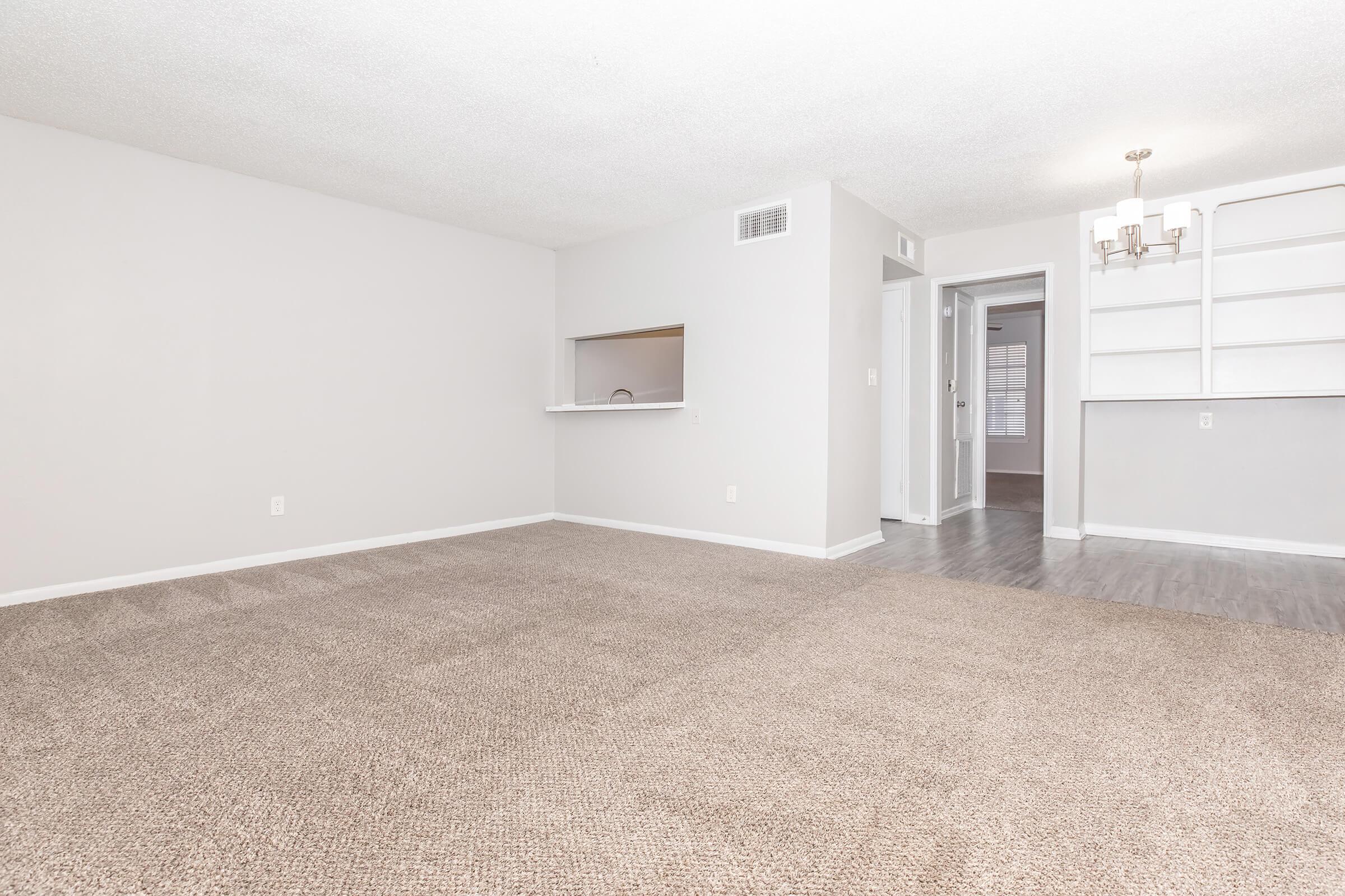 Spacious, empty living room with light beige carpet and neutral-colored walls. A small pass-through opens to a kitchen area. To the right, a doorway leads to another room or hallway. A chandelier hangs from the ceiling, and there's a built-in shelf on the far wall, adding to the room's natural light.