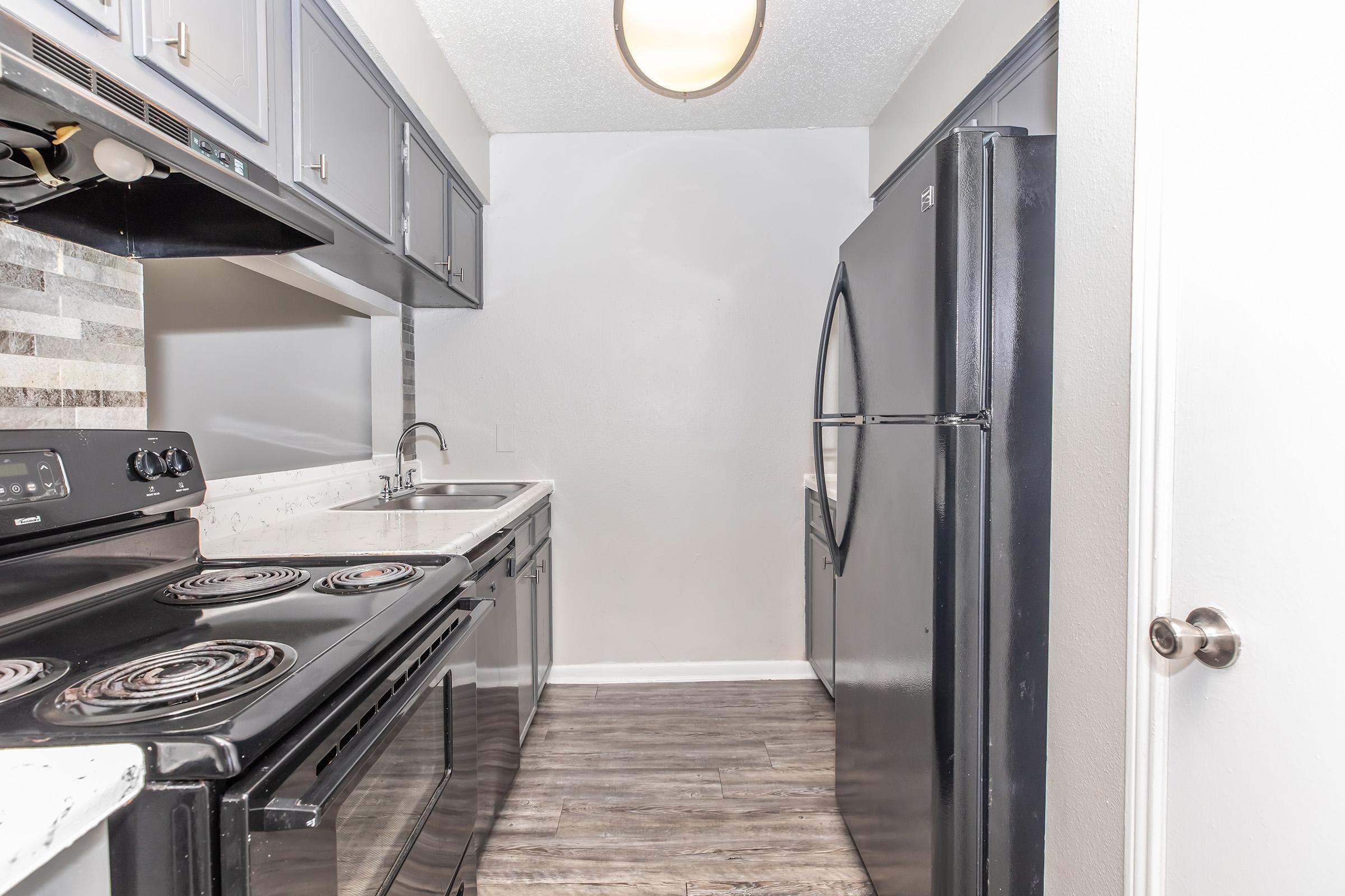 View of a modern kitchen featuring dark cabinets, a stainless steel sink, an electric stove, and a black refrigerator. The walls are painted light gray, and there is a decorative backsplash above the countertop. The floor has a wood-like finish, adding warmth to the space.