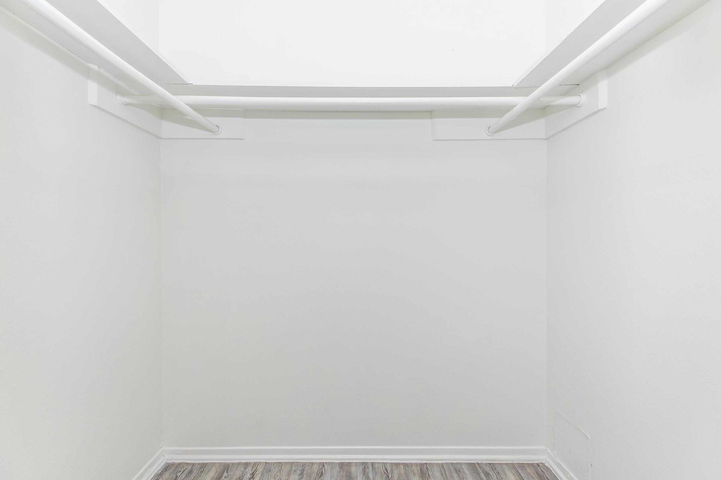 Empty closet with white walls and two horizontal hanging rods. The floor has a light wood-like finish, and there is no visible clutter or items inside the space.
